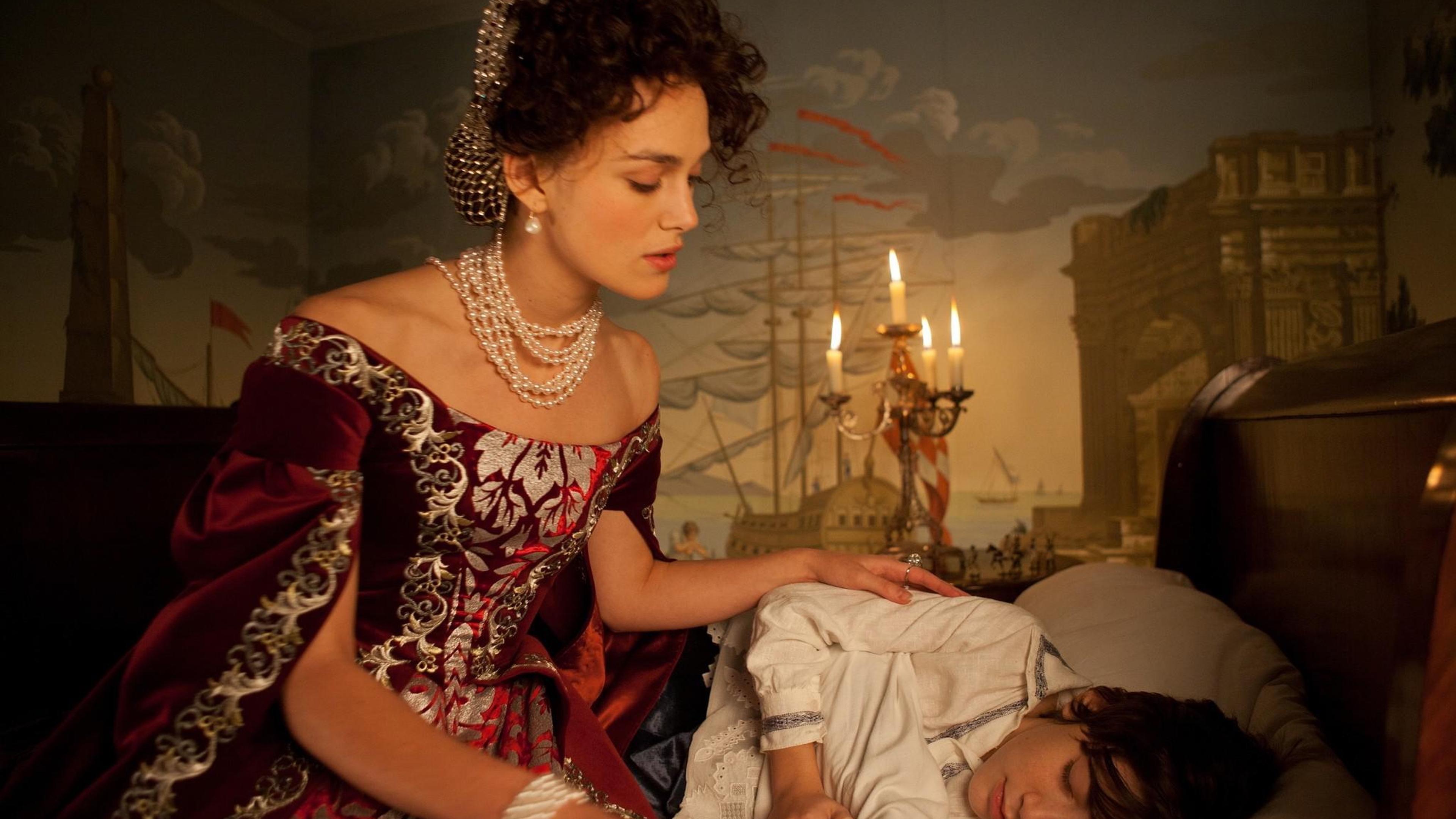 A photo of a woman in historical attire watching over a sleeping child by candlelight, with a mural of ships in the background.
