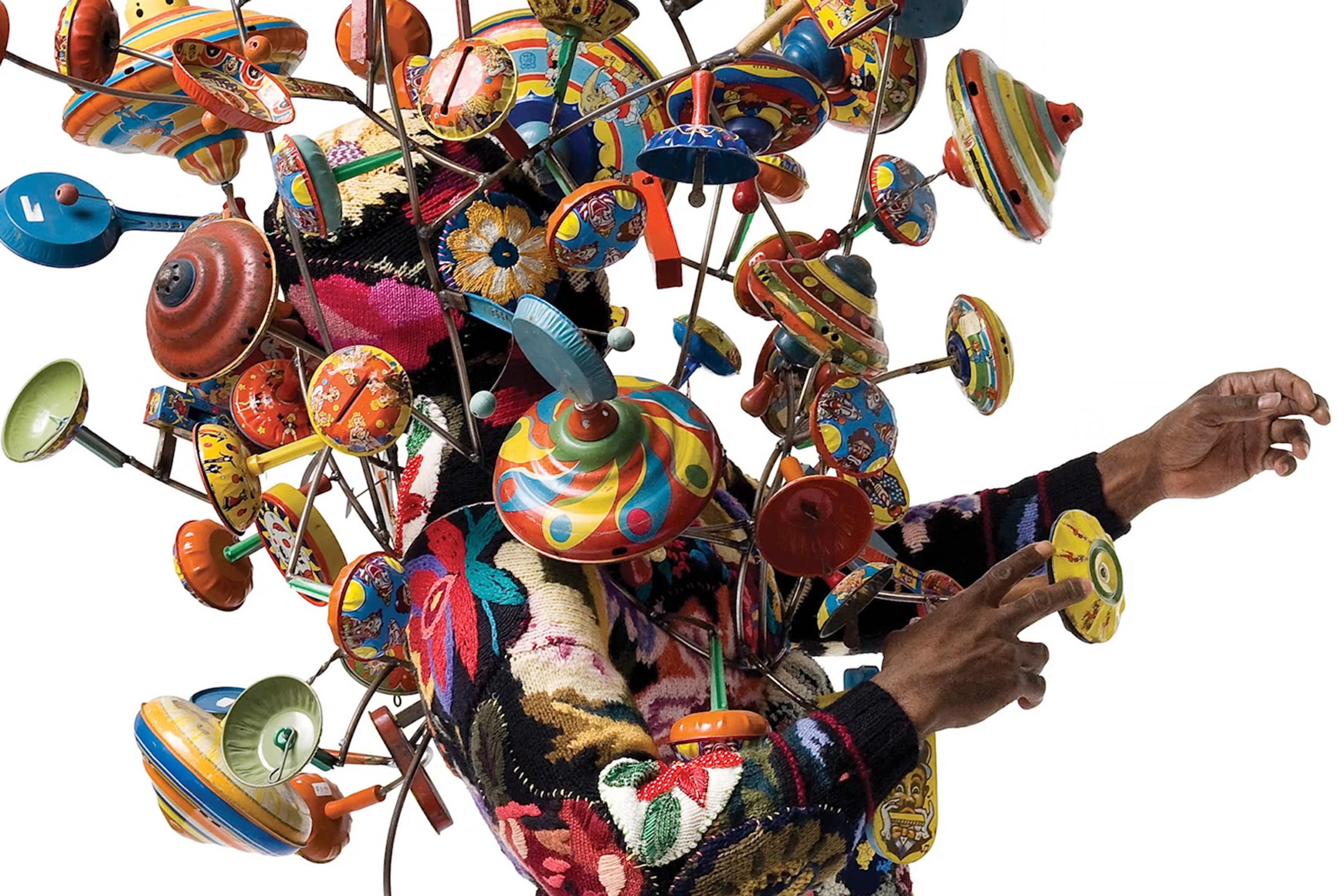 A person wearing a colourful patterned sweater, surrounded by numerous vibrant, vintage spinning tops on metal rods.