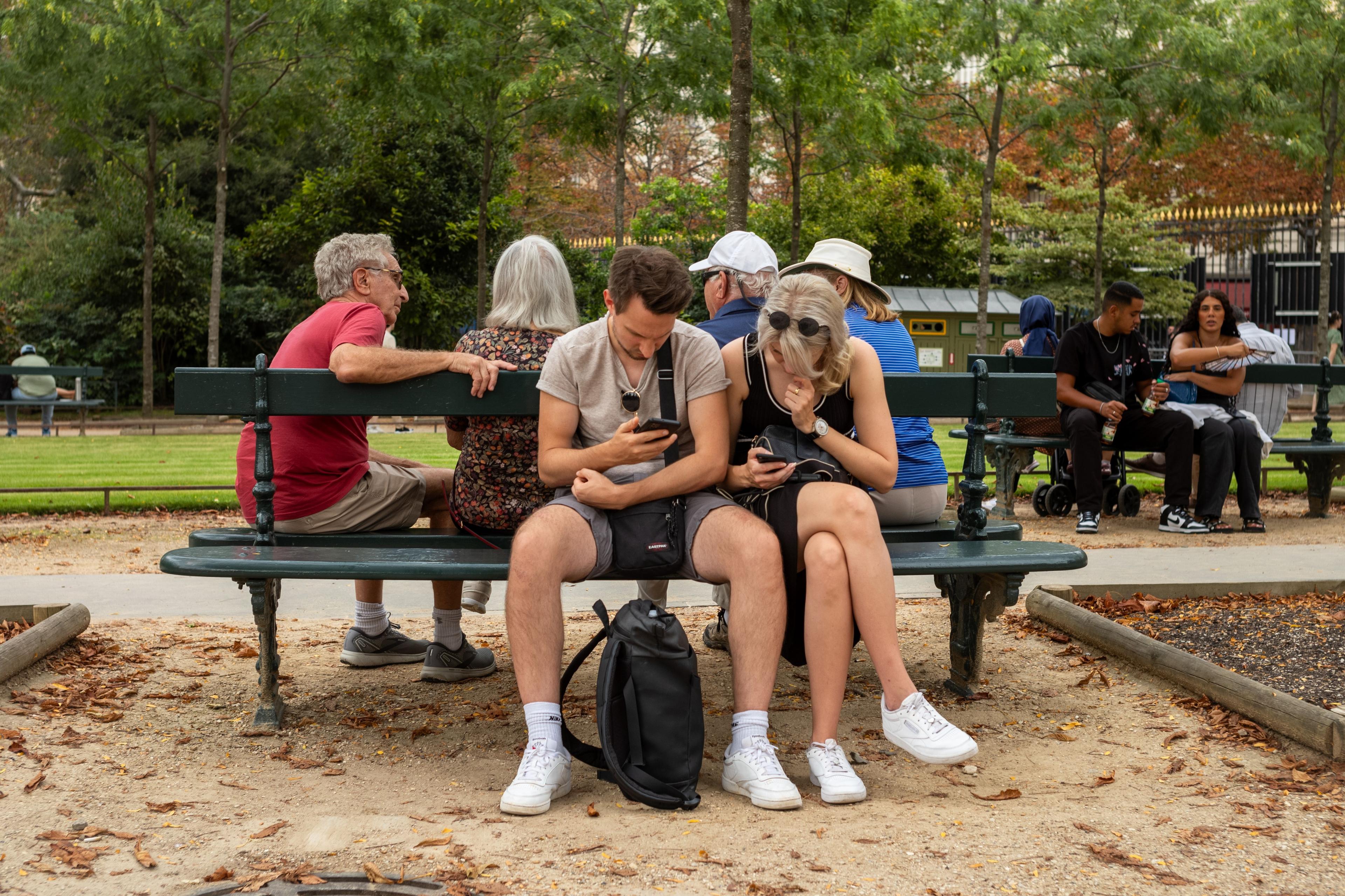 Photo of people sitting on a park bench looking at phones, surrounded by trees and other park visitors.