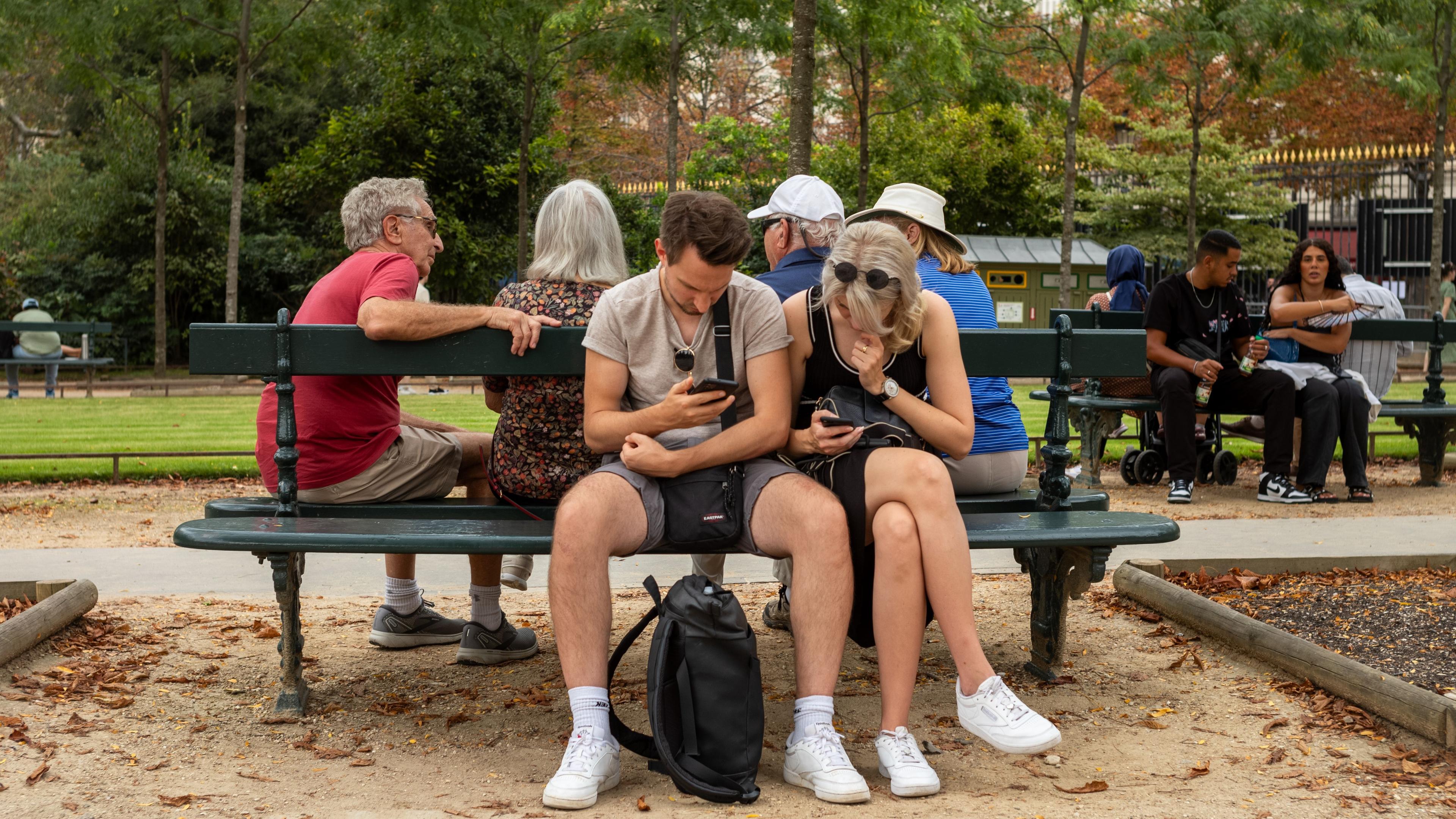Photo of people sitting on a park bench looking at phones, surrounded by trees and other park visitors.