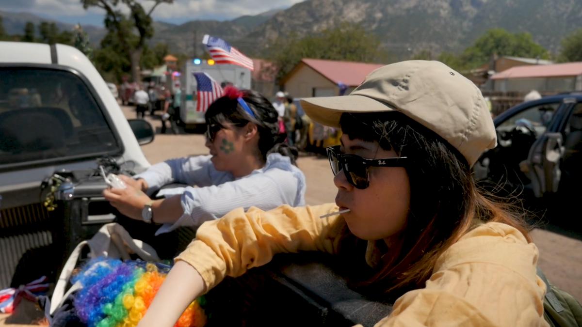 Photo of two young Asian women wearing sunglasses sitting in the back of a pickup truck at an outdoor event with American flags and mountains.