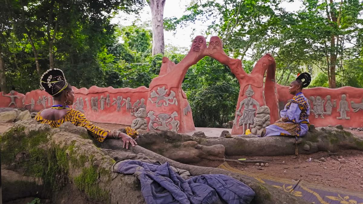 Photo of two women in traditional attire sitting by a painted sculpture in a lush forest setting.