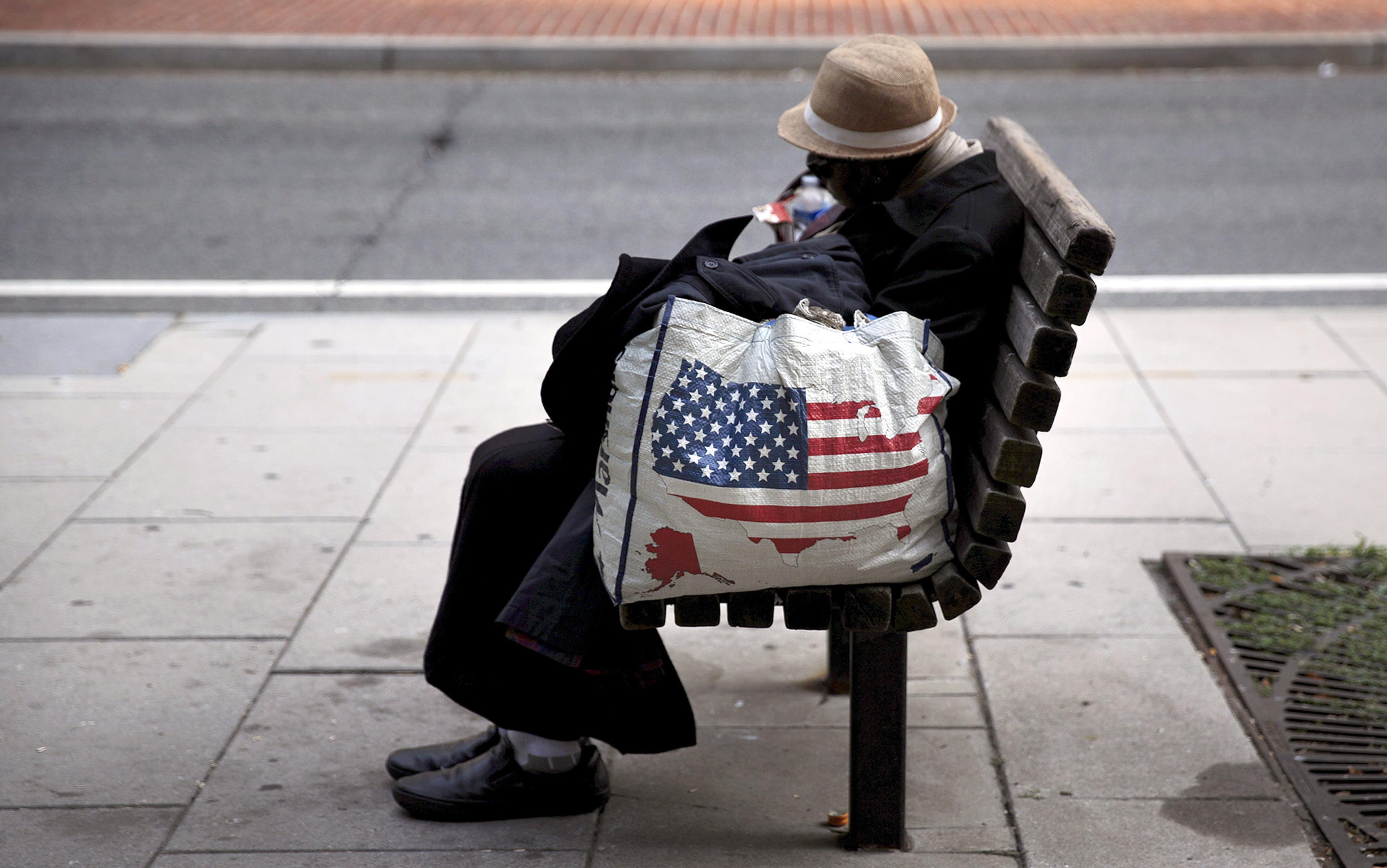 A man in a hat sitting asleep on a bench, holding a shopping bag with an American flag design on a city pavement.