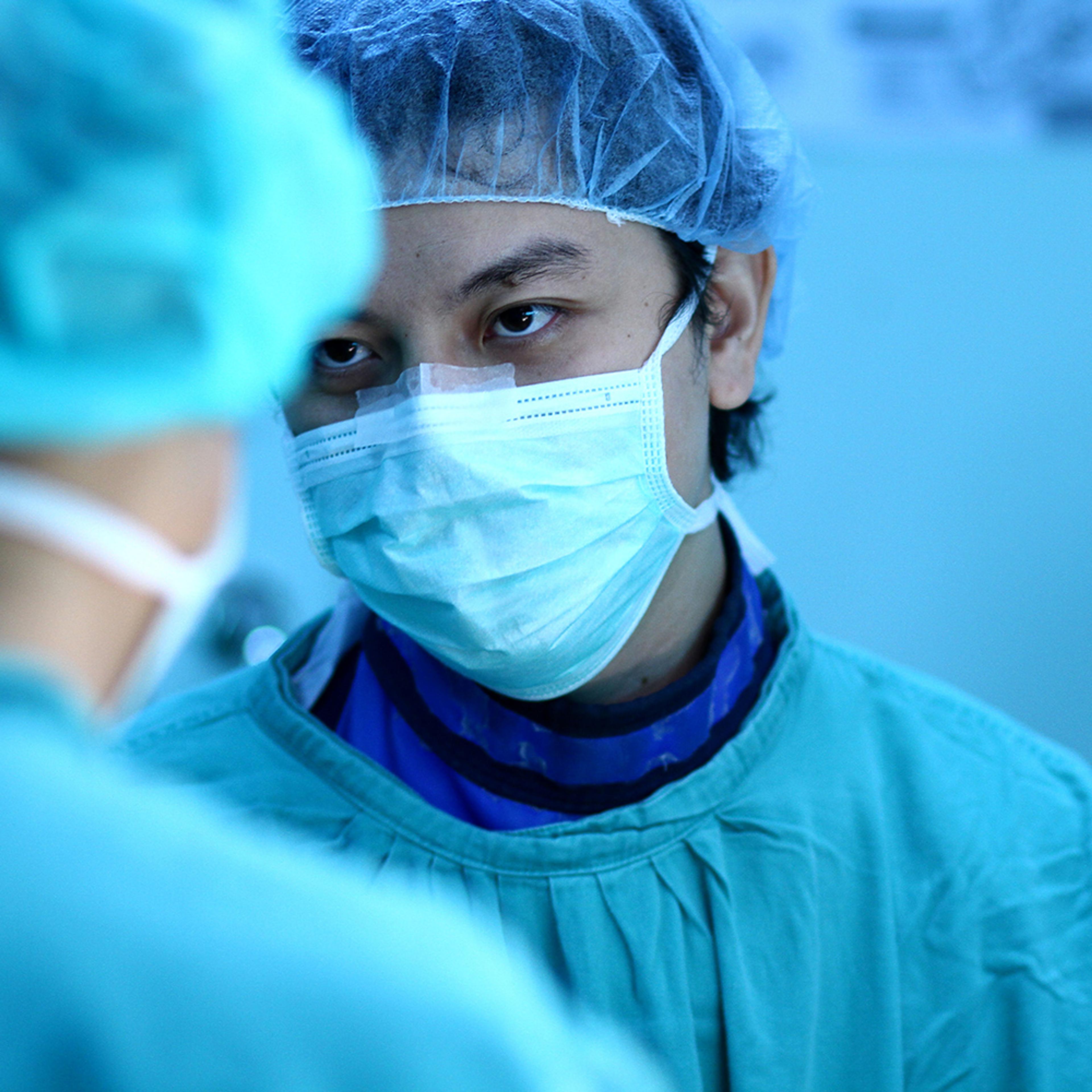 A surgeon in a surgical mask, blue scrubs and a hairnet, focused intently on a colleague in a hospital setting.