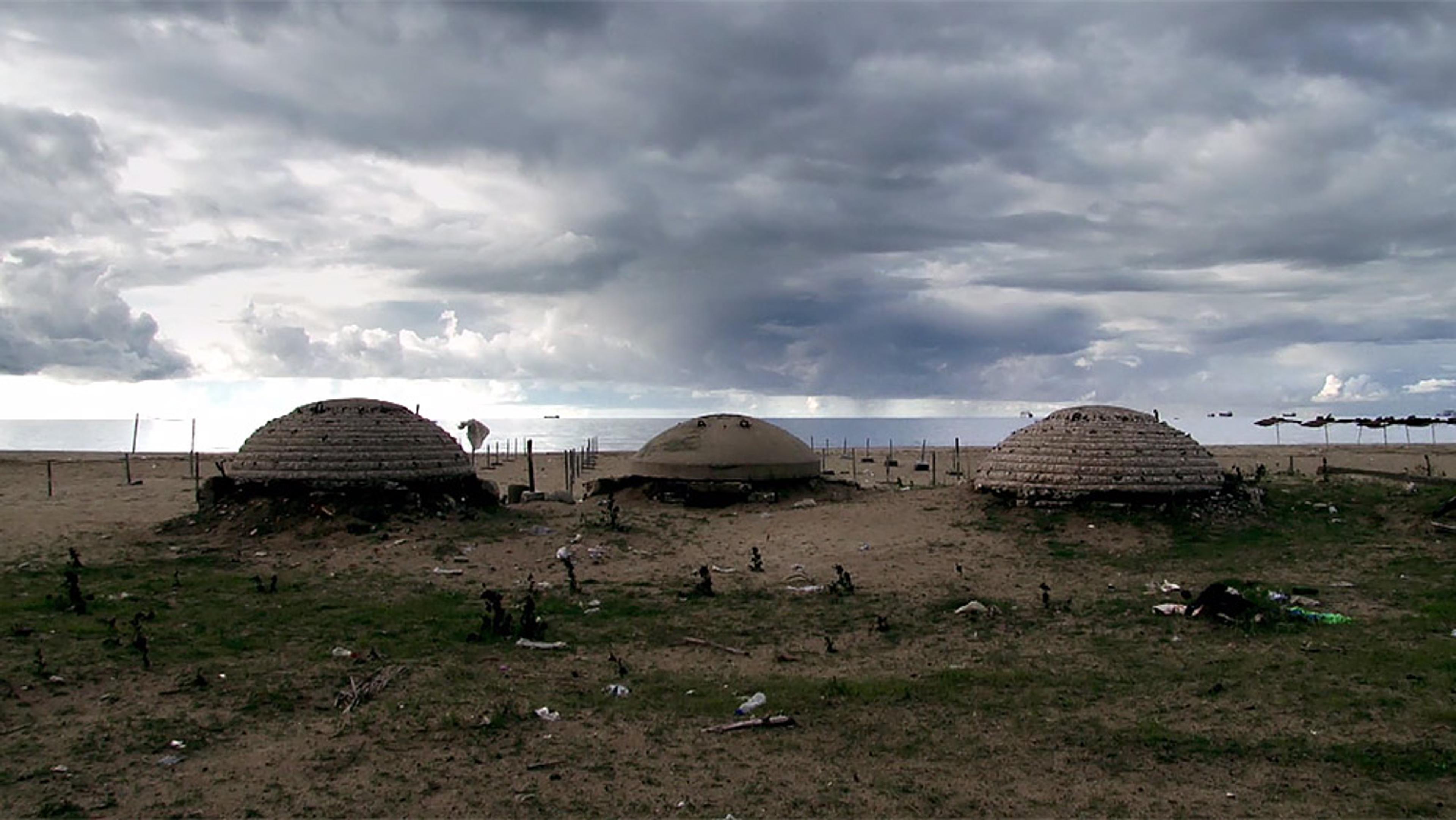 Three dome-shaped bunkers on a beach with a cloudy sky, bits of trash on the ground, and the sea in the background.