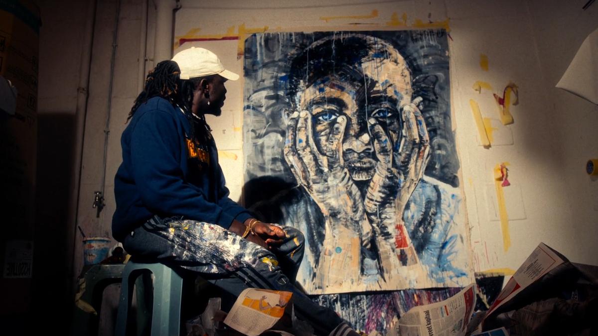 A photo of an artist in a cap and paint-splattered trousers sitting next to a large painting of a man holding his face in his hands.