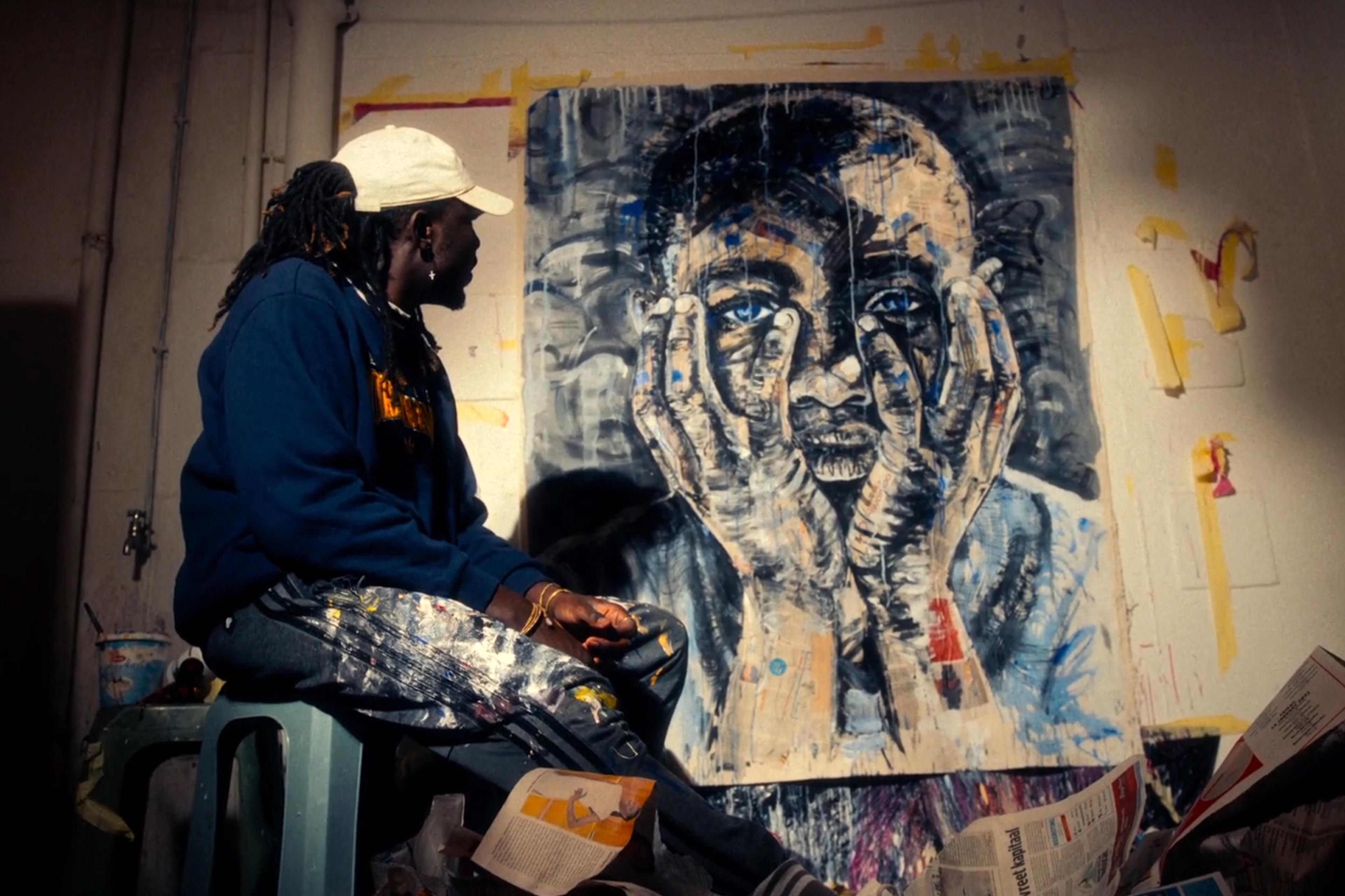 A photo of an artist in a cap and paint-splattered trousers sitting next to a large painting of a man holding his face in his hands.