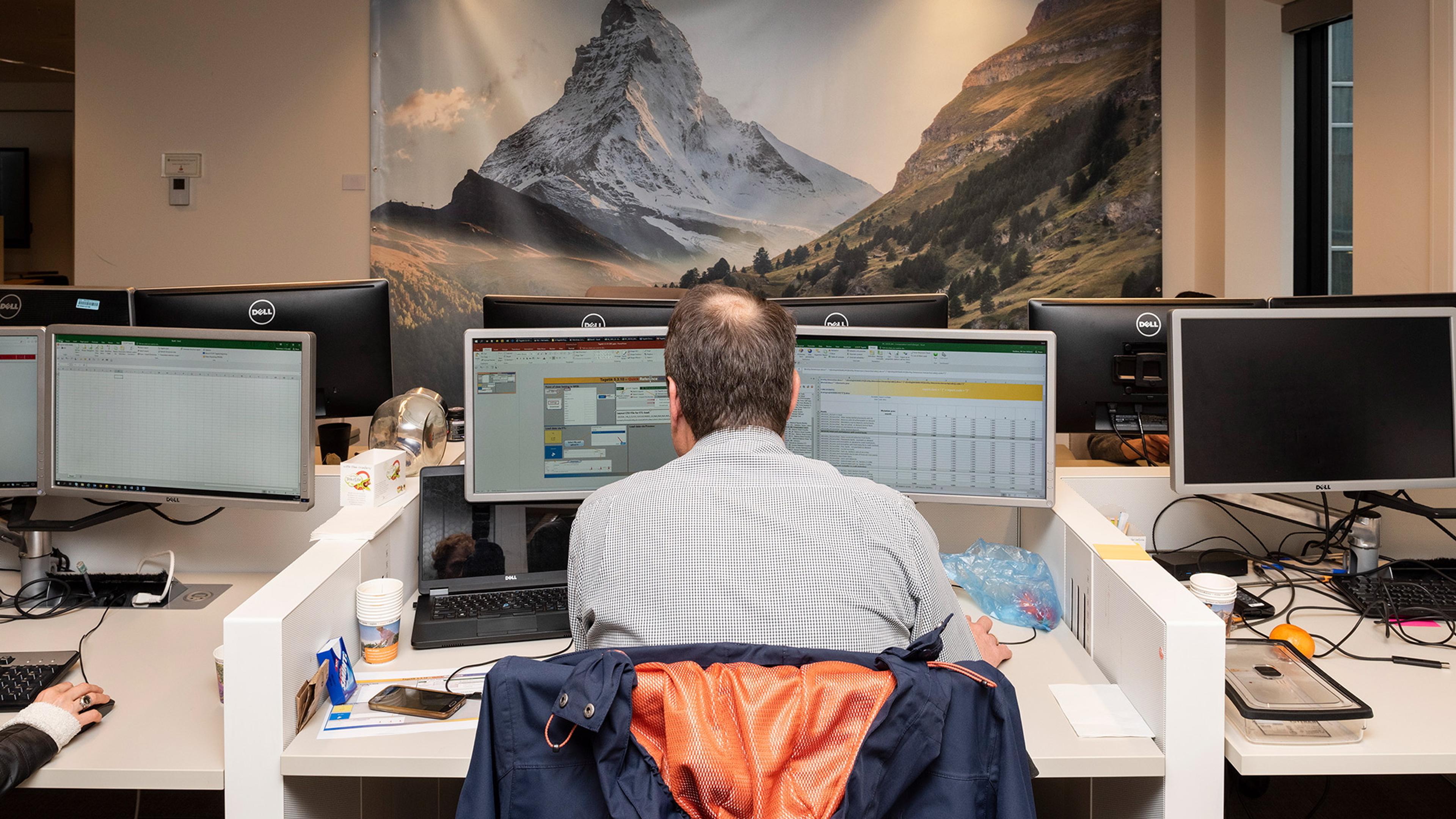 Photo of a man working at a desk with multiple monitors, backed by a large mountain mural, in an office setting.