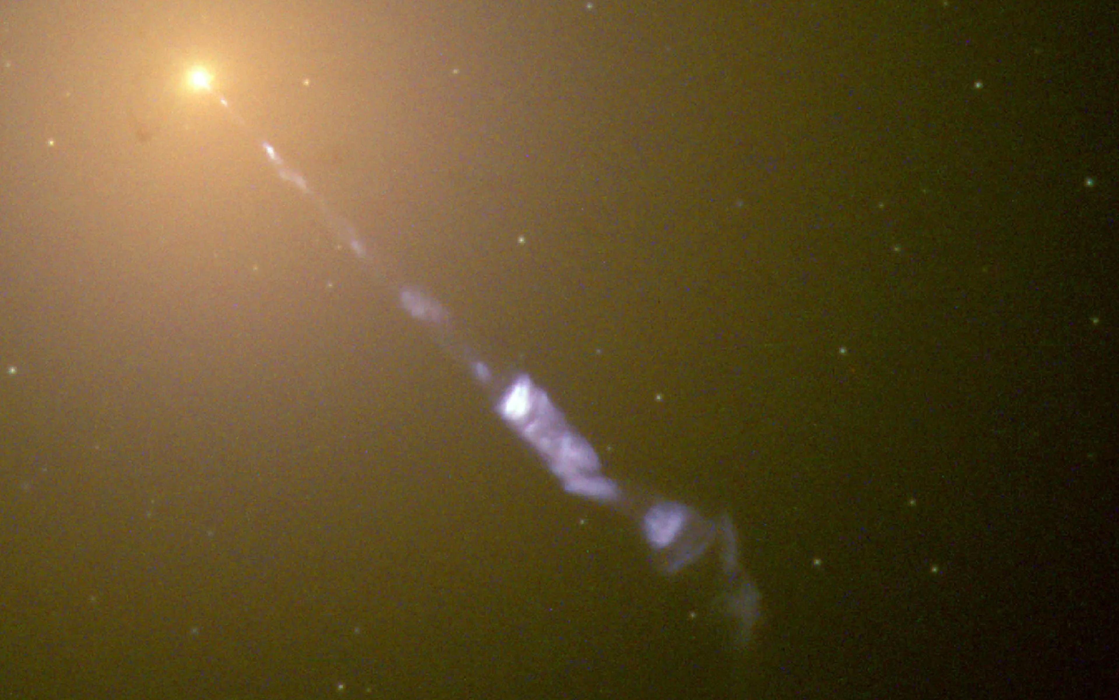 Image of M87 galaxy showing a bright yellowish central core with a jet of blue plasma extending outward into space. The background is filled with faint stars and a hazy, brownish hue