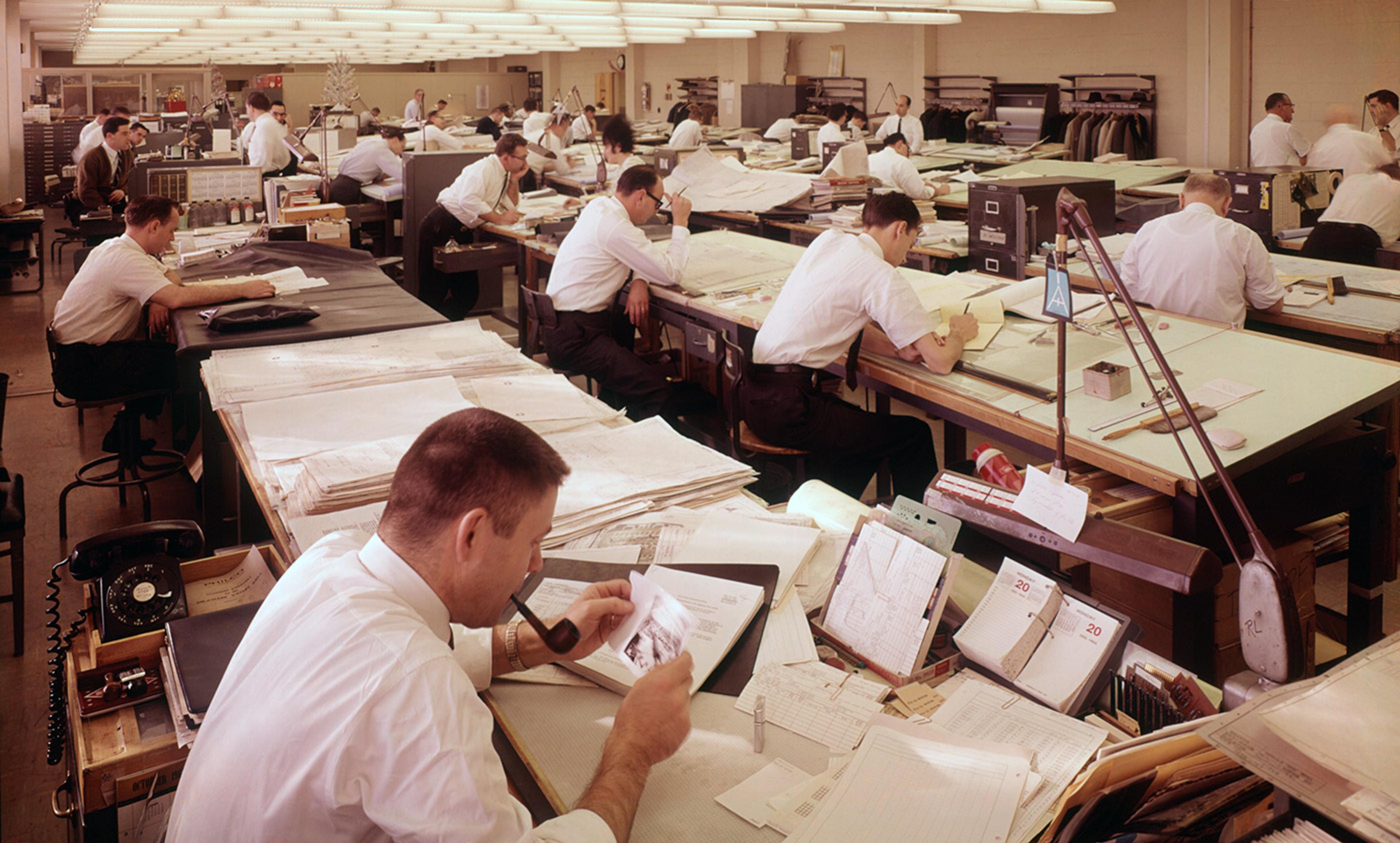Vintage photo of a large 1960s office with men at drafting tables working on papers and designs under bright overhead lights.