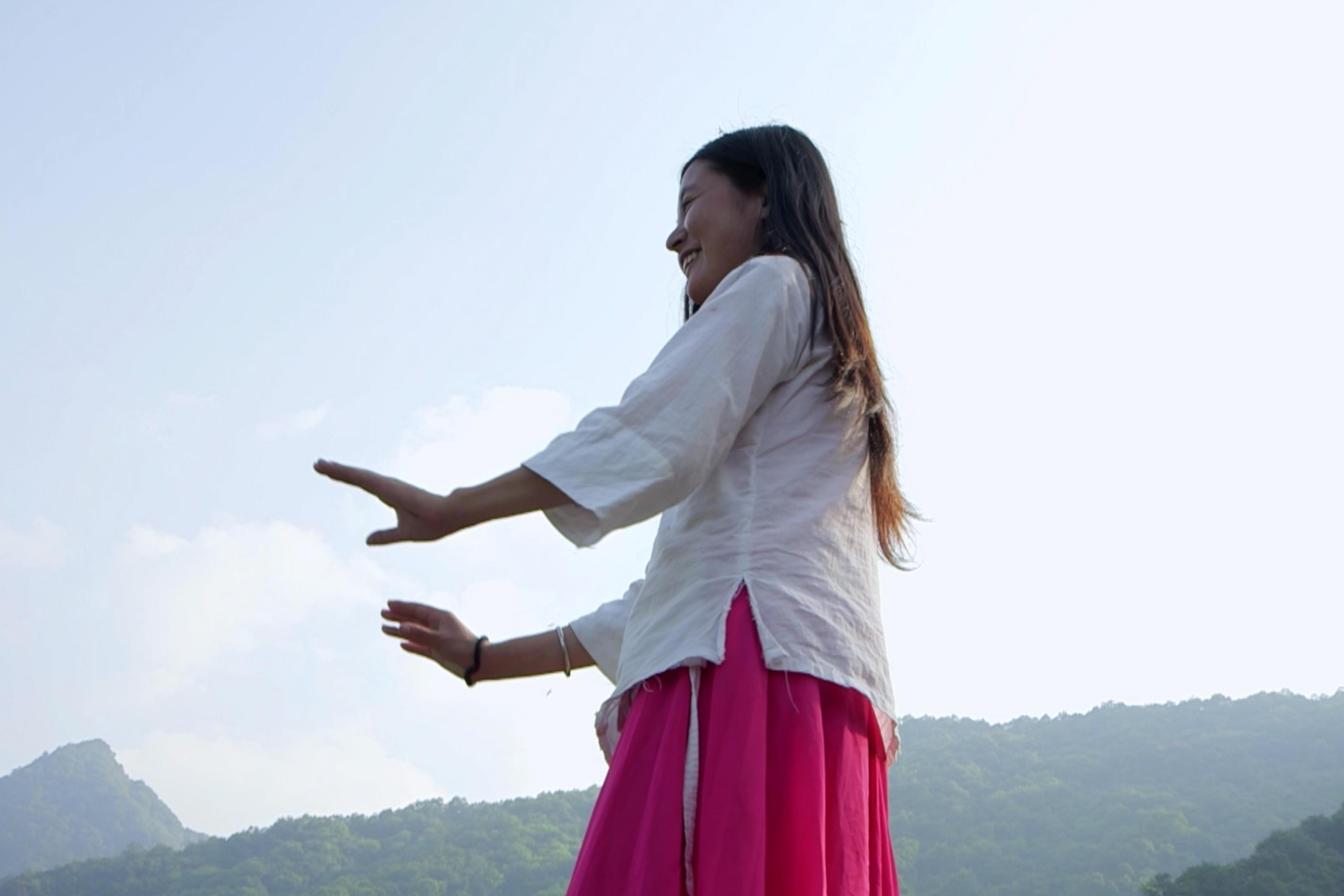 A Chinese woman with long hair wearing a white top and pink skirt with arms outstretched against a backdrop of mountains and sky.