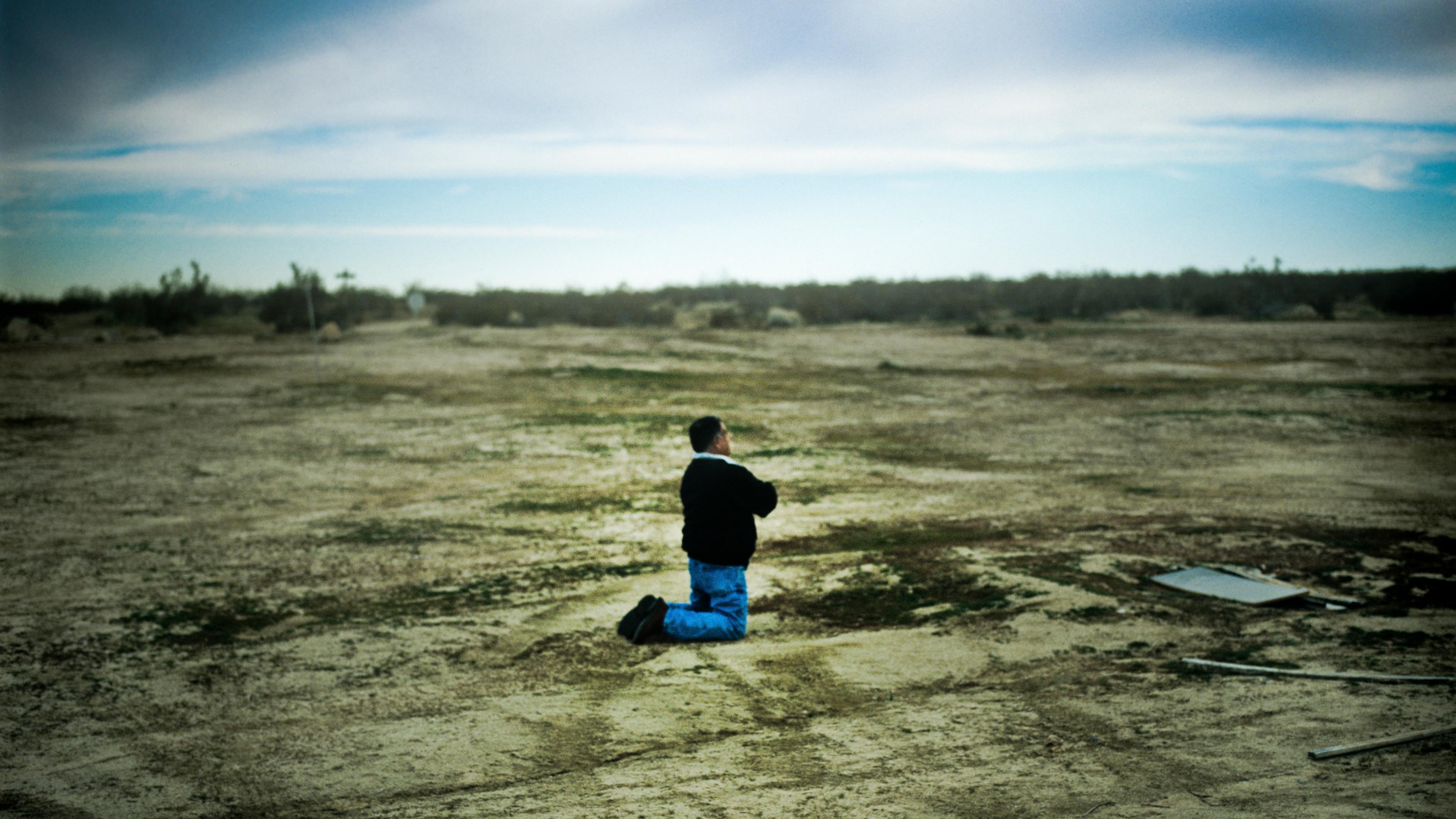 Photo of a person kneeling on a barren landscape under a cloudy sky, capturing a sense of isolation and vastness.