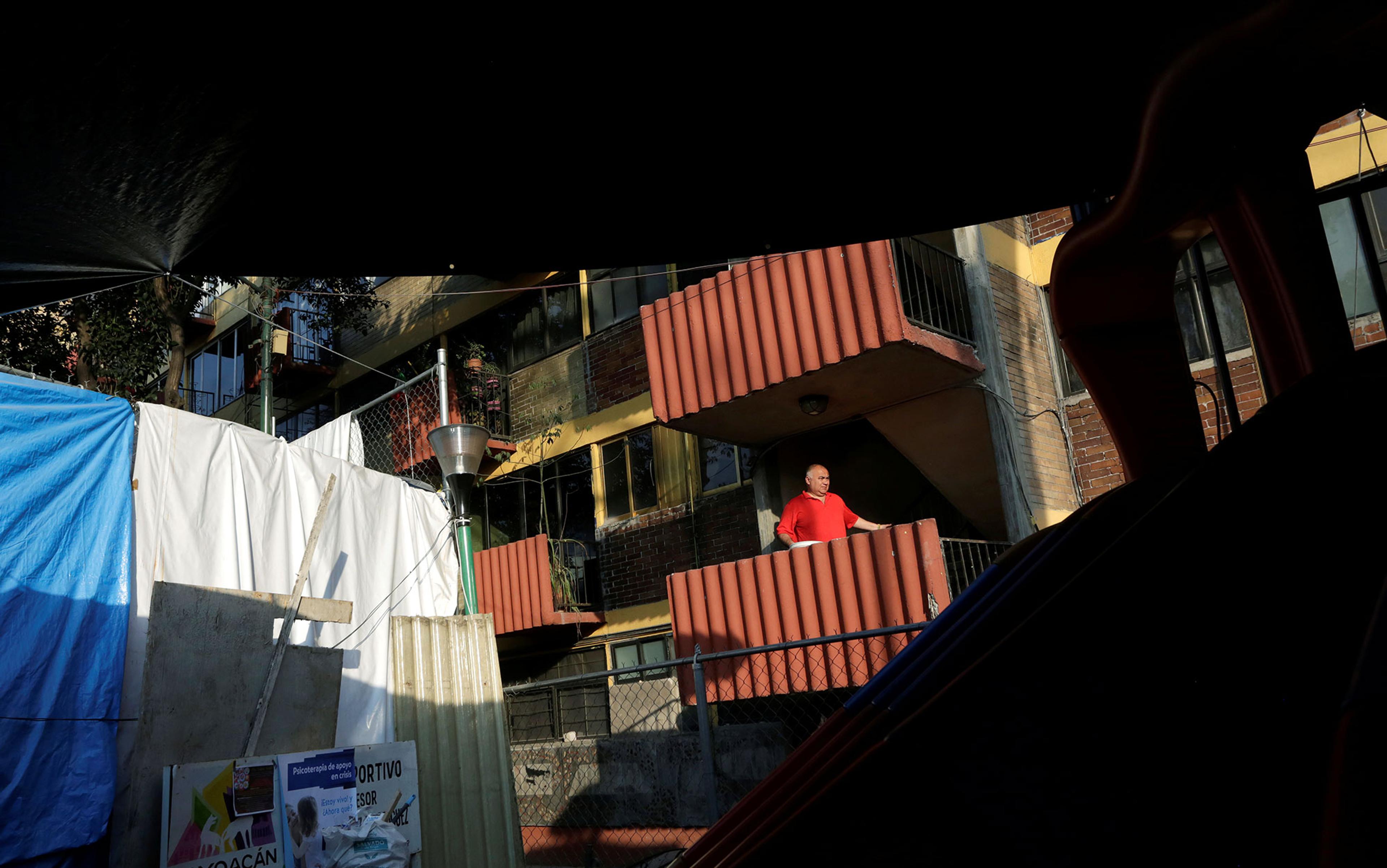 A man standing on a balcony of a brick apartment building, with tarps and debris in foreground.