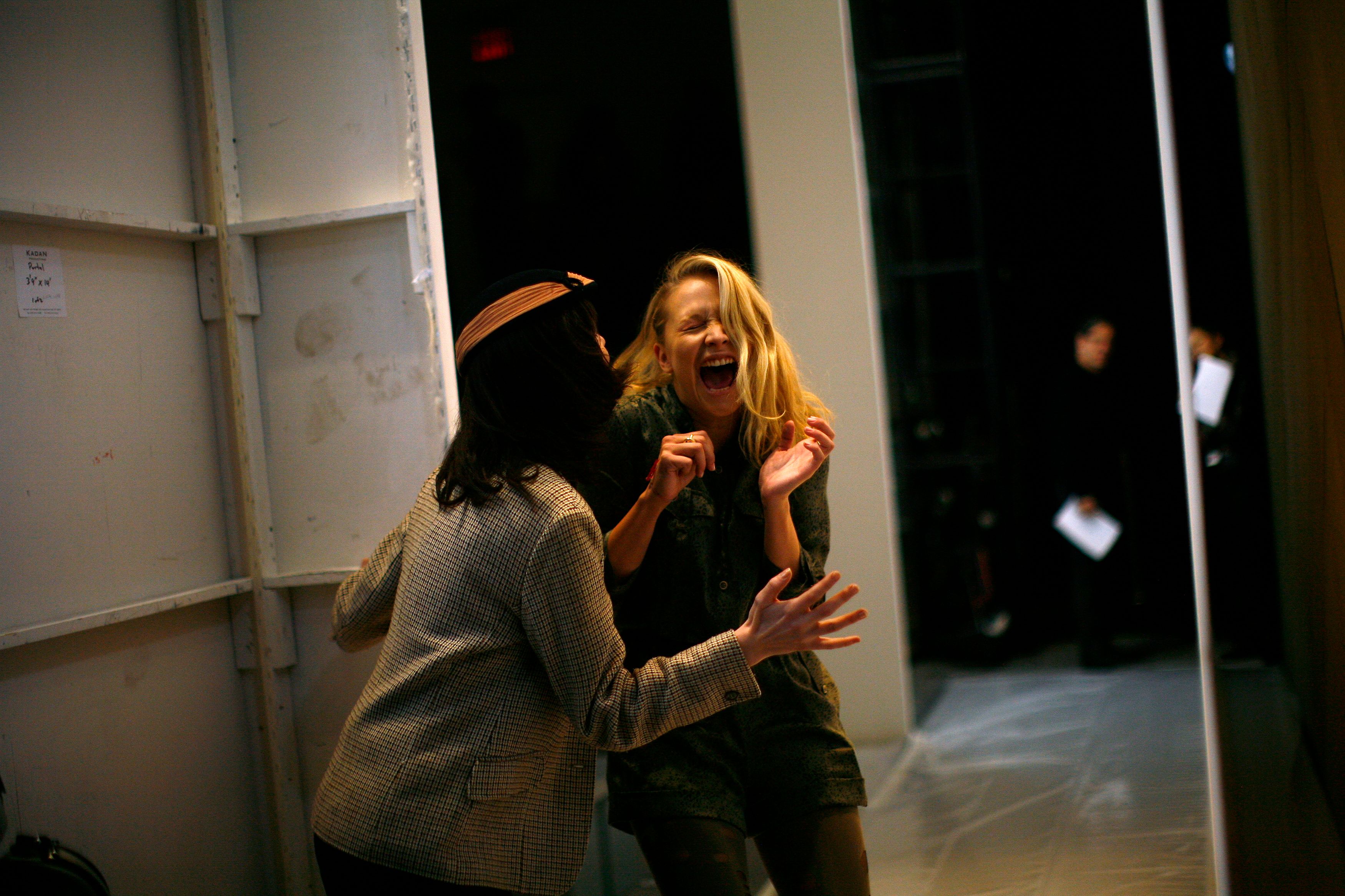 Photo of two women laughing backstage, one facing away and the other facing forward, with blurred figures in the background.