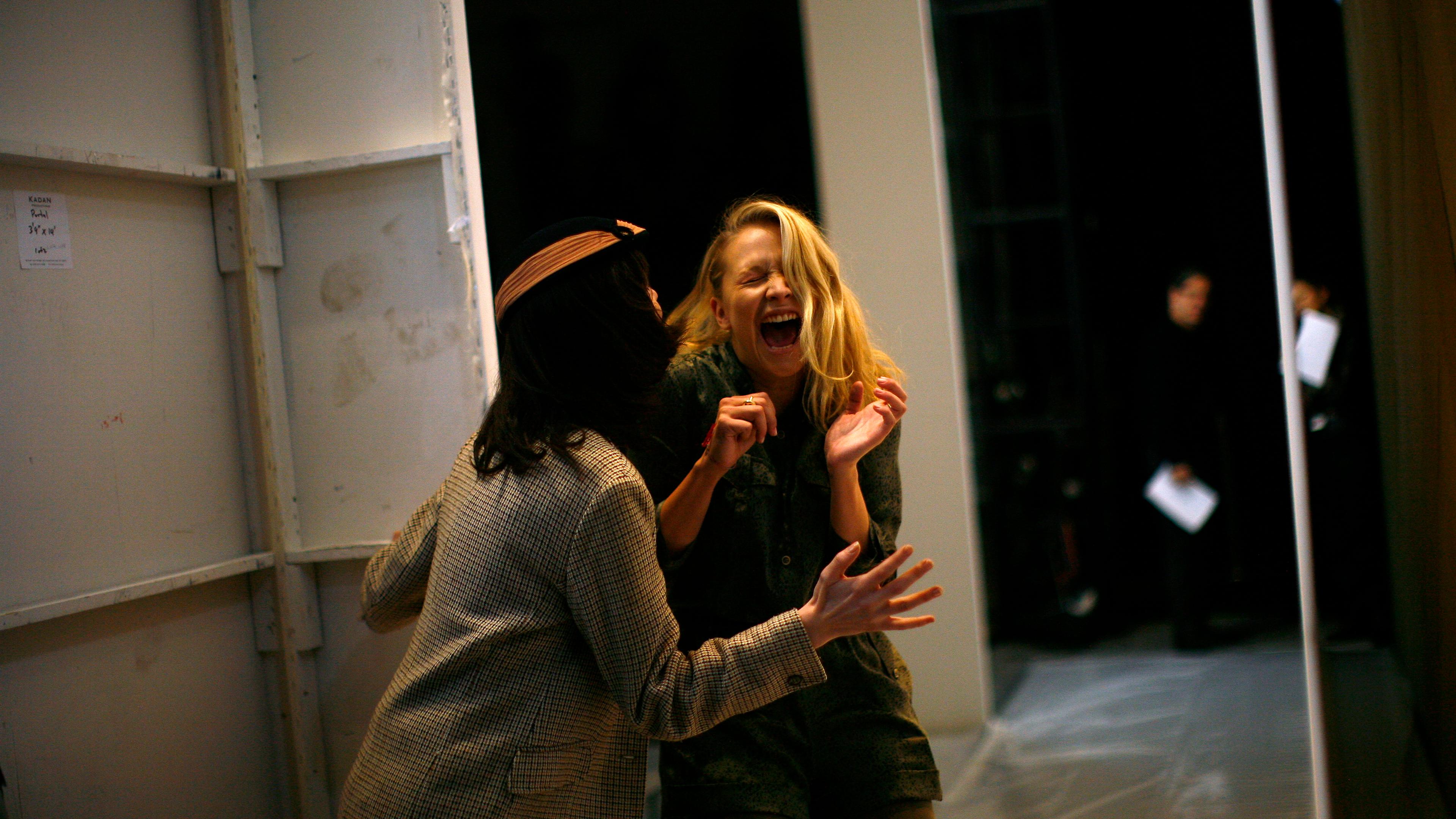 Photo of two women laughing backstage, one facing away and the other facing forward, with blurred figures in the background.