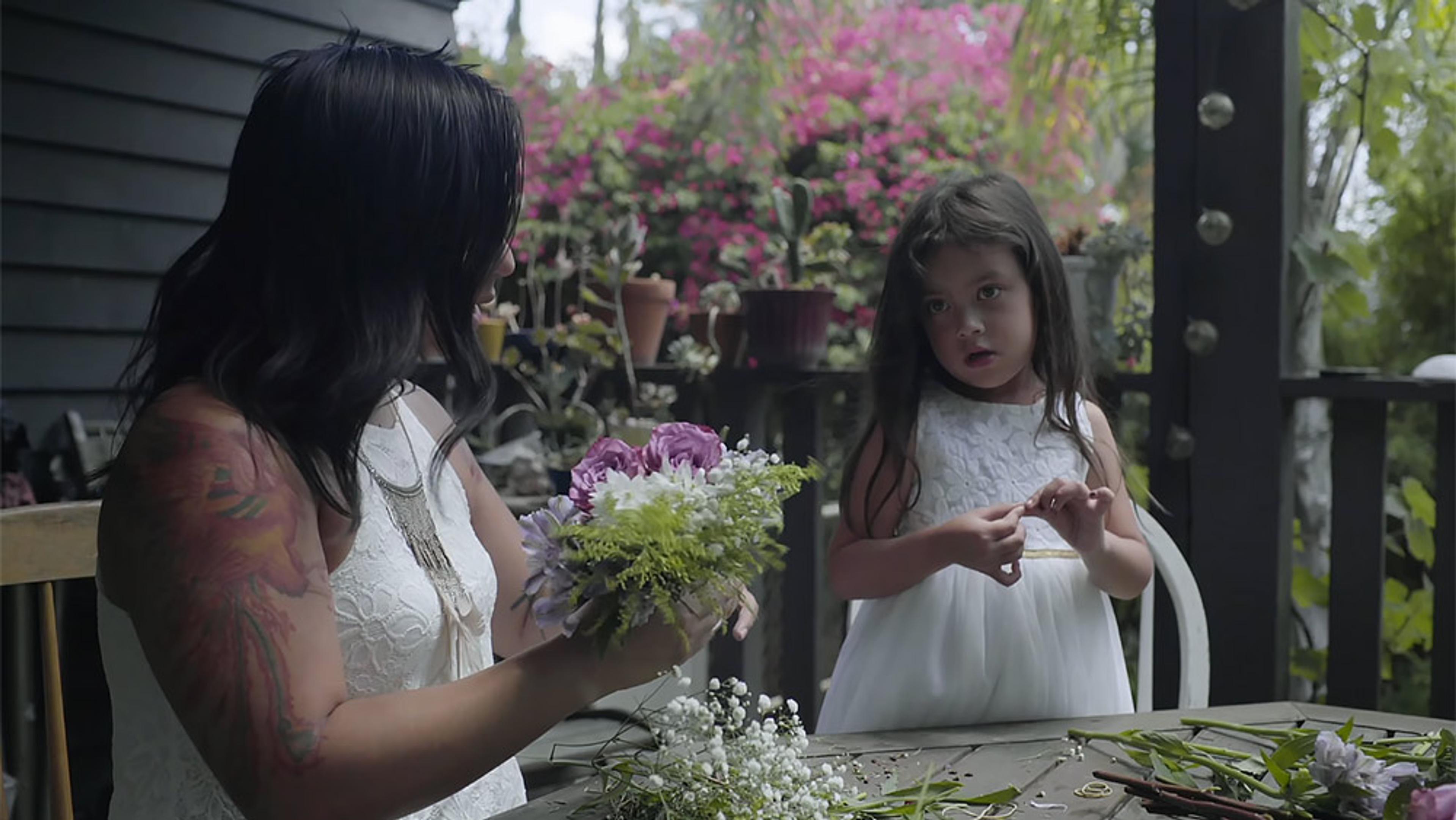 A young woman and a little girl both with long dark hair and in white dresses arranging flowers at a table on a porch.