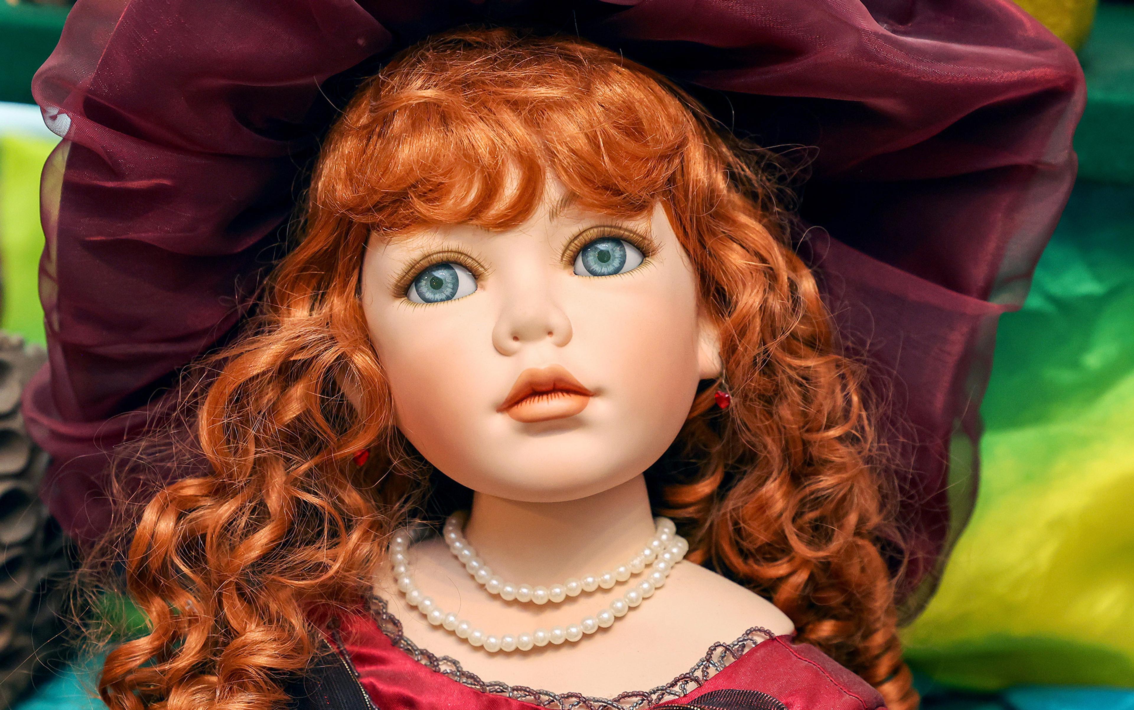 Photo of a doll with curly red hair, blue eyes, and a large maroon hat, wearing a double string of pearls and a red dress.