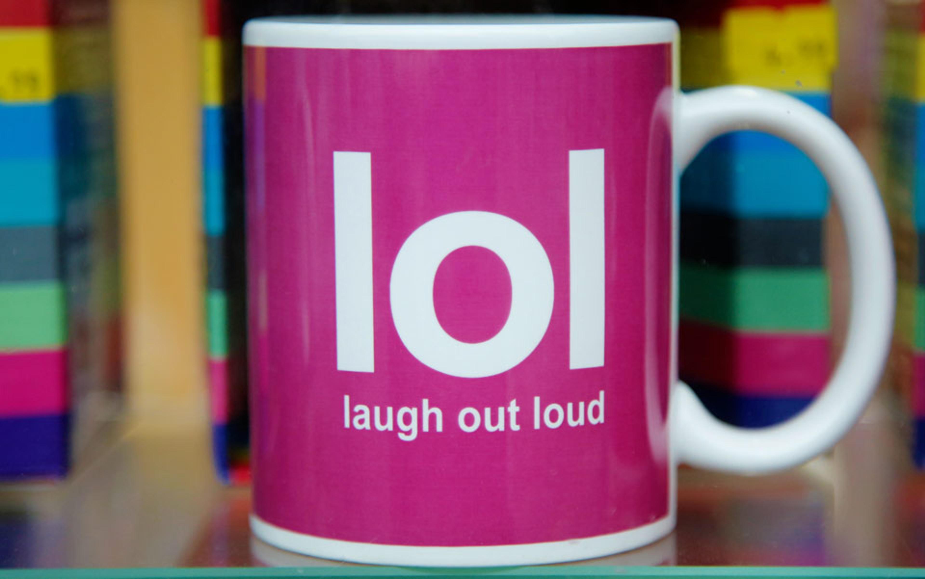 A pink mug with “lol laugh out loud” printed in white. The background features colourful, stacked objects.