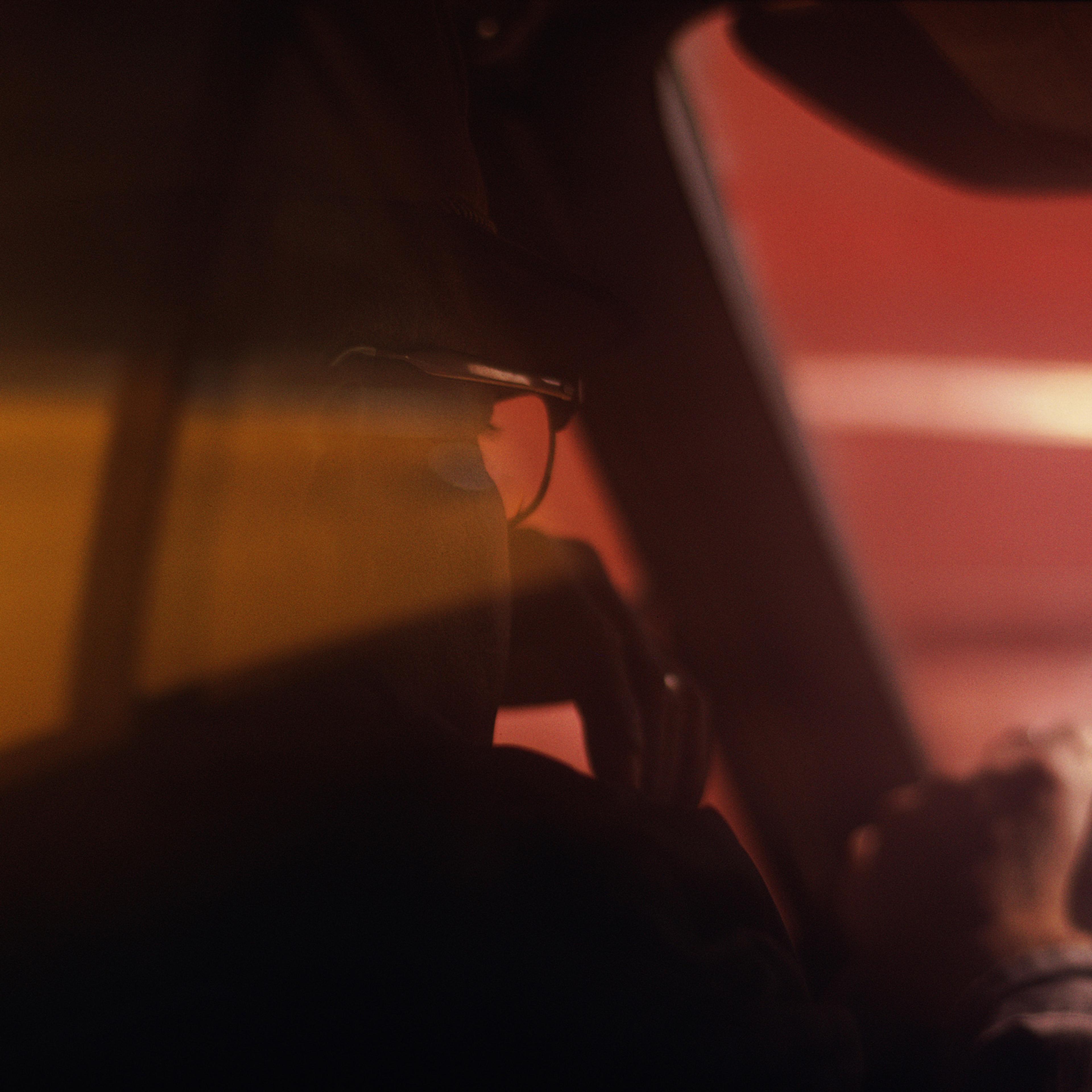 Photo of a person driving a car, seen through a car window, lit by warm sunlight, wearing glasses, with one hand on the steering wheel and the other hand resting in front of their face.