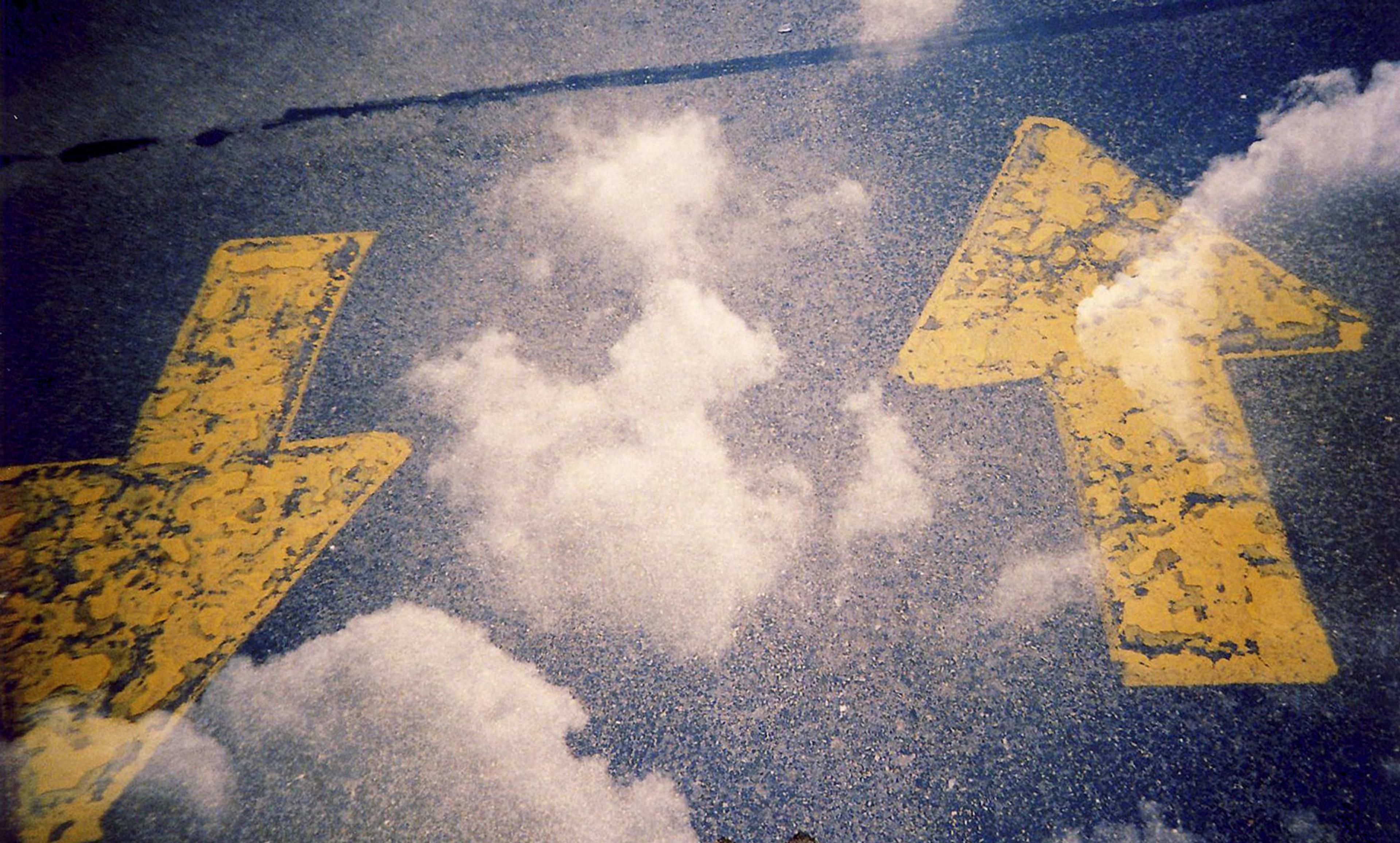 Two yellow road arrows painted on asphalt, pointing left and right, with a reflection of clouds on the surface.