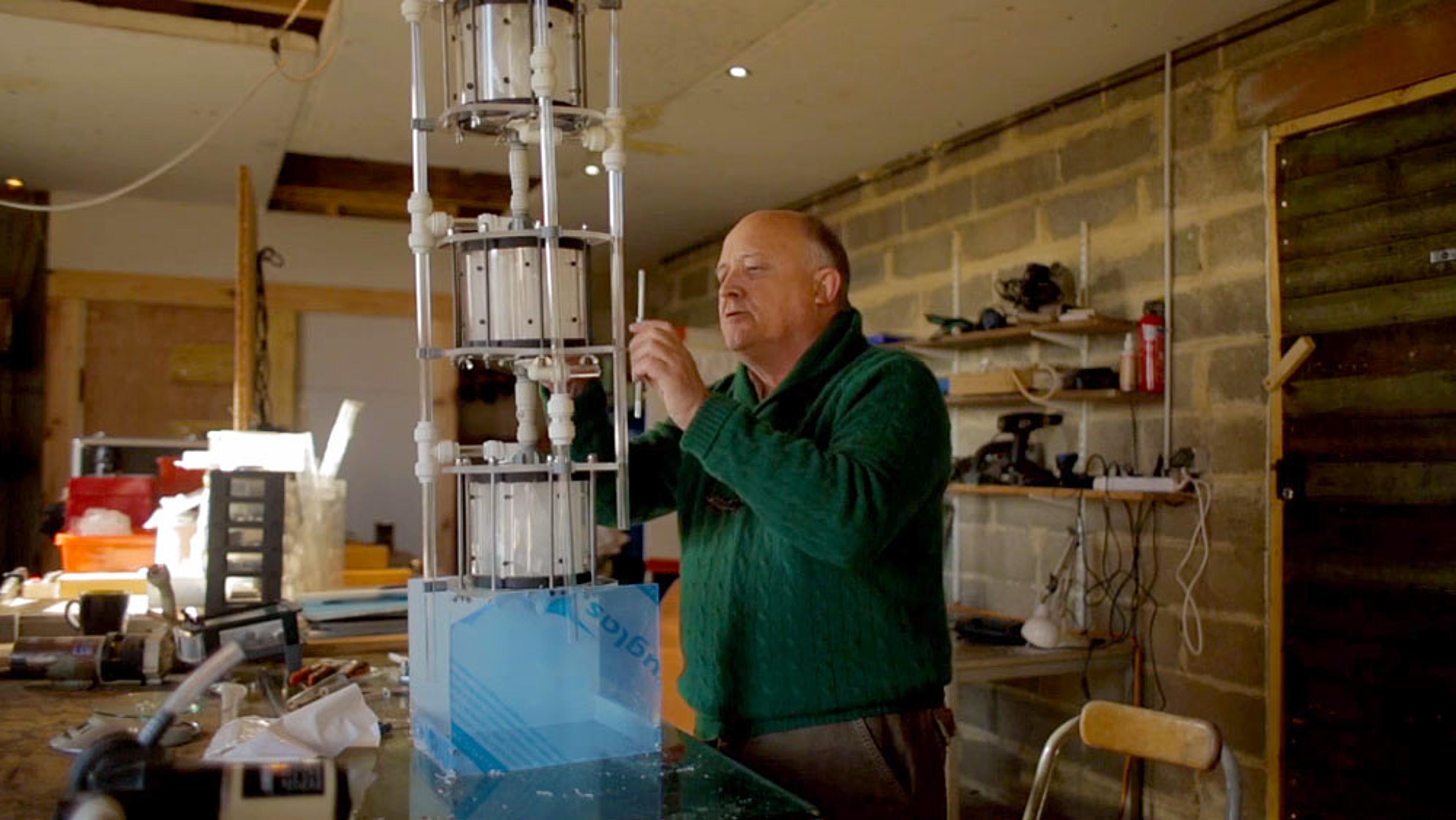 A man in a green jumper working on a vertical mechanical device in a cluttered workshop, with wooden shelves and tools.