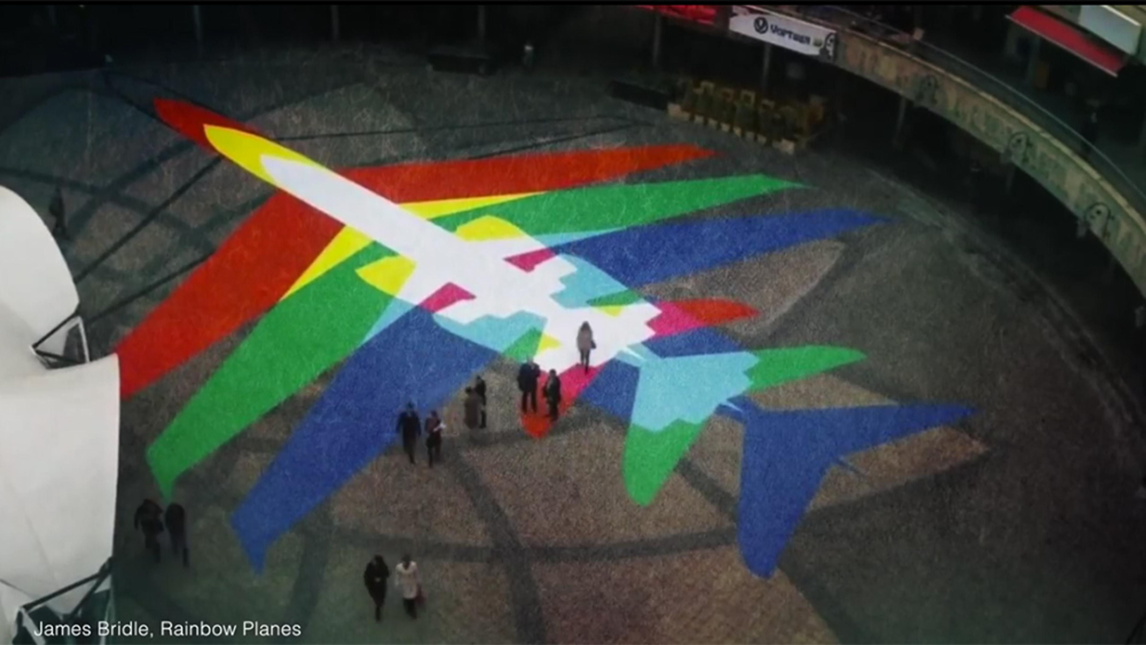 Aerial photo of a colourful plane mural on a public square with people walking around it. Text: James Bridle, Rainbow Planes.