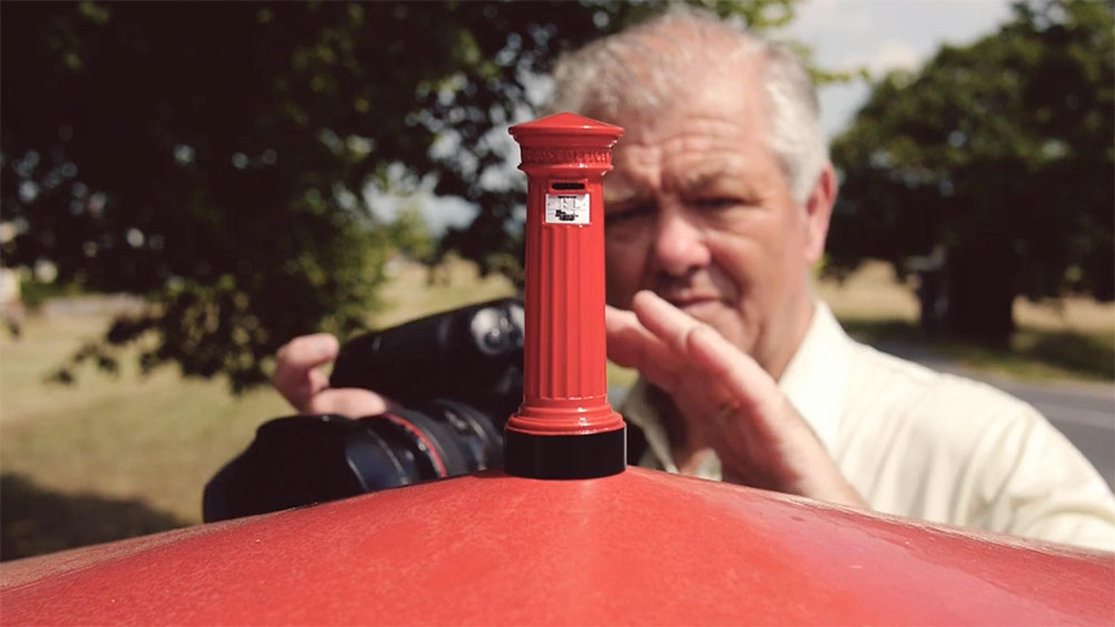 A man taking a picture of a miniature British red postbox on top of a regular sized postbox, with a blurred background of trees and sunlight.