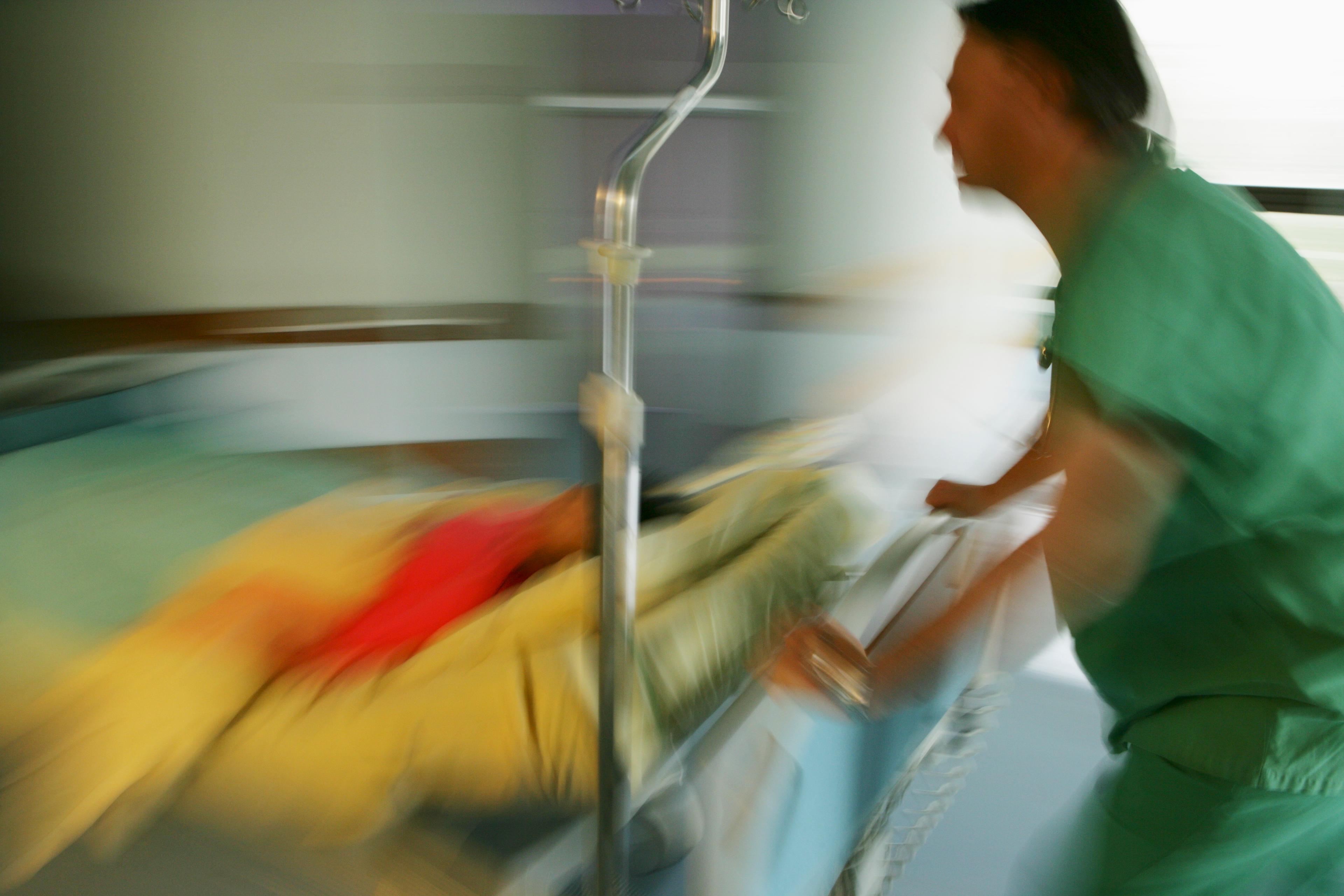 Blurred photo of a nurse in green uniform pushing a hospital gurney with a patient in a yellow blanket for urgent care.