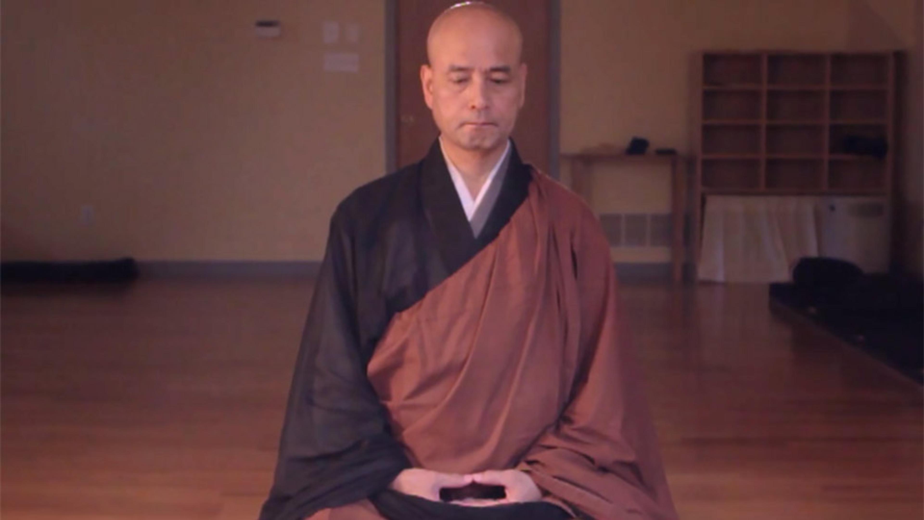 A bald man in traditional robes sitting on the floor meditating in a simple room with wooden floors and minimal furniture.