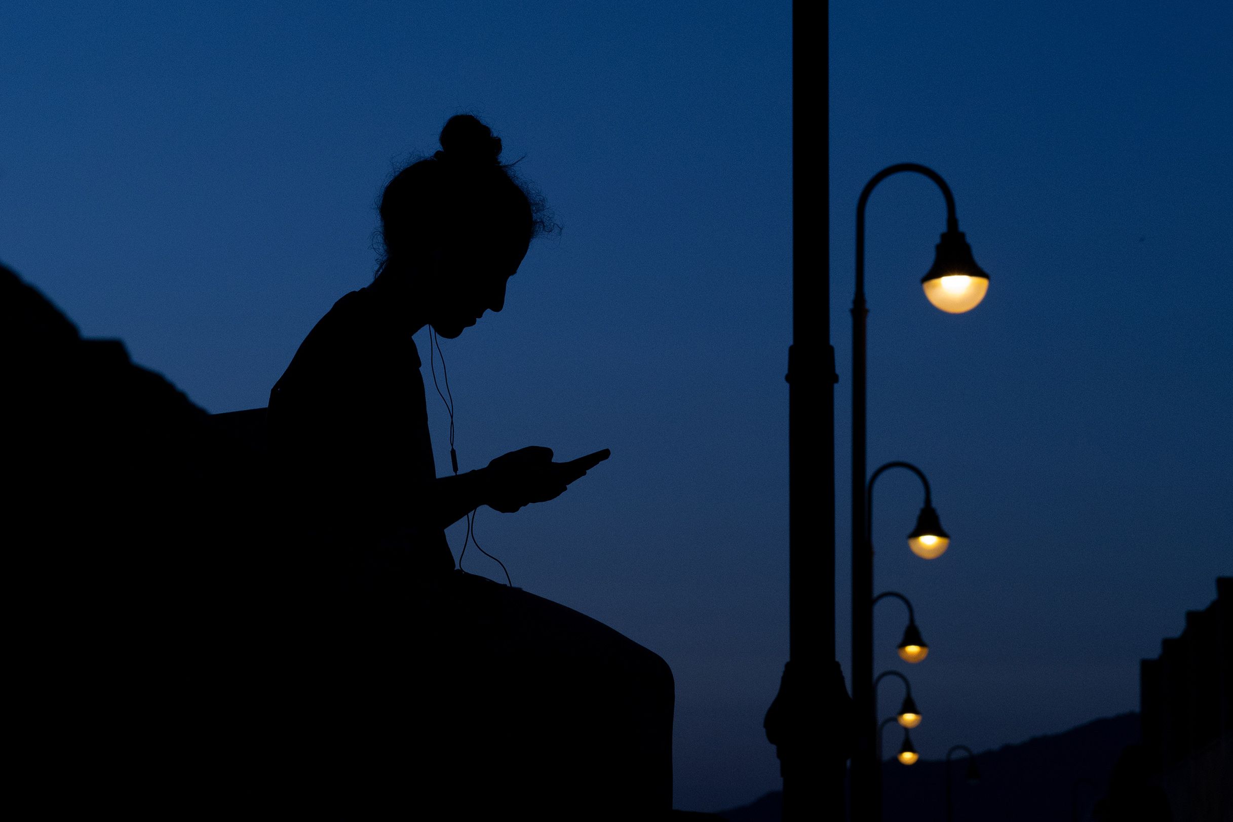 Silhouette of a person with headphones holding a phone against a twilight sky with streetlamps in the background.