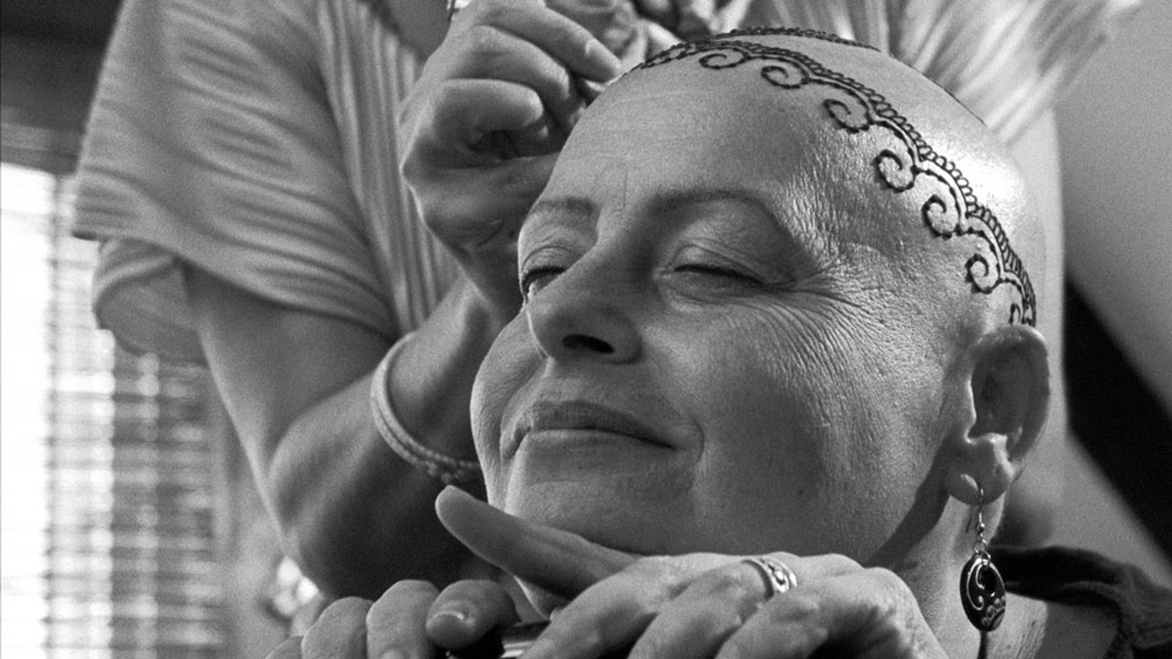 A black and white photo of a smiling bald woman getting a decorative tattoo or henna pattern on her head by another person.