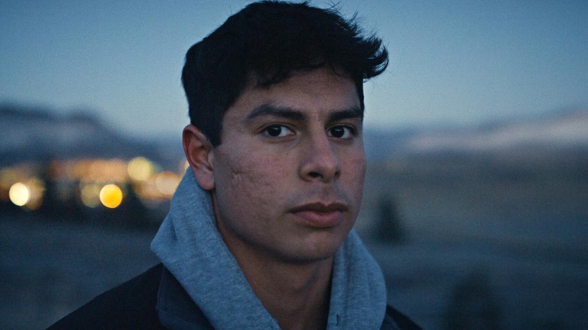 A young man with a serious expression wearing a grey hoodie outdoors at dusk, with a blurred landscape and lights in the background.