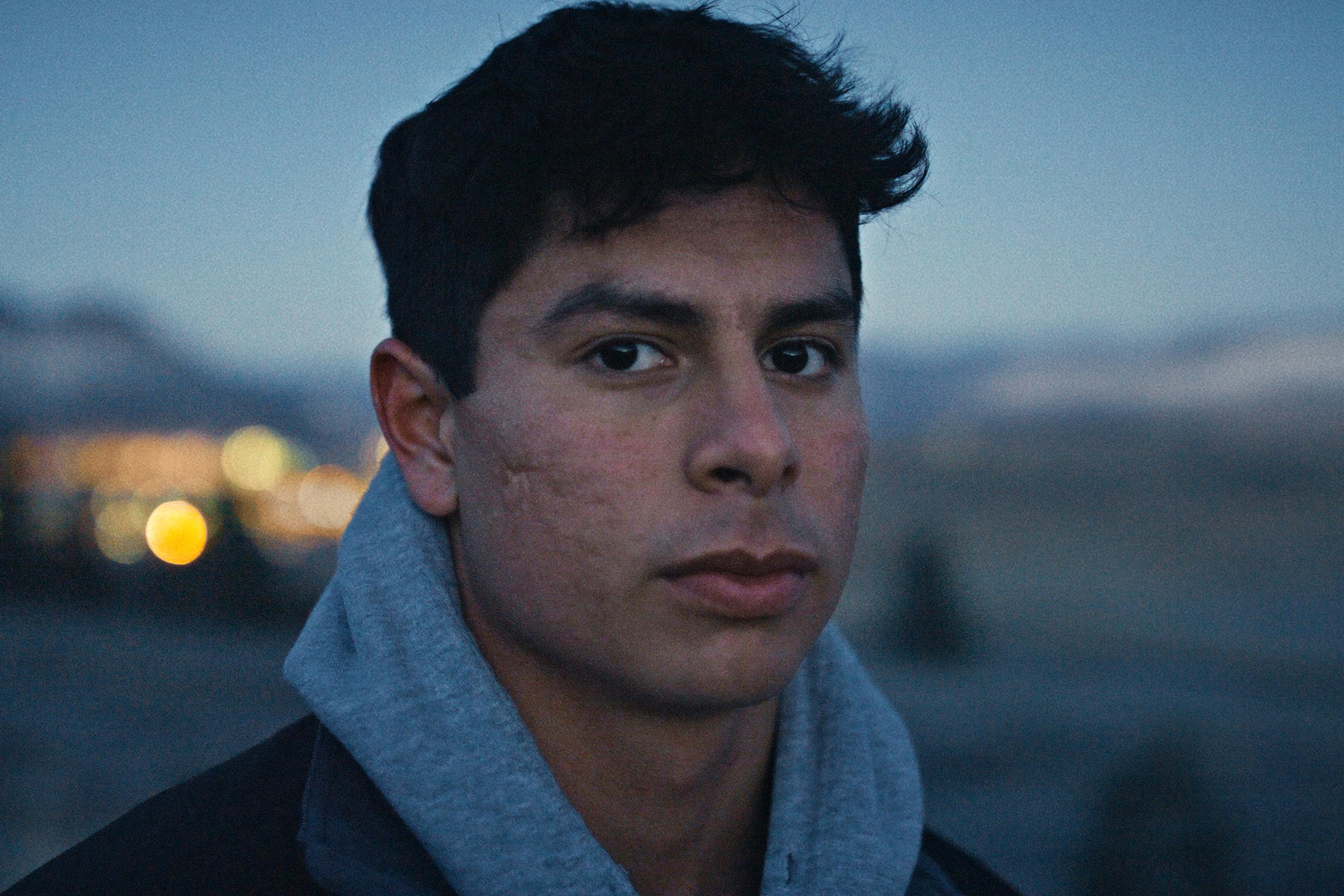 A young man with a serious expression wearing a grey hoodie outdoors at dusk, with a blurred landscape and lights in the background.