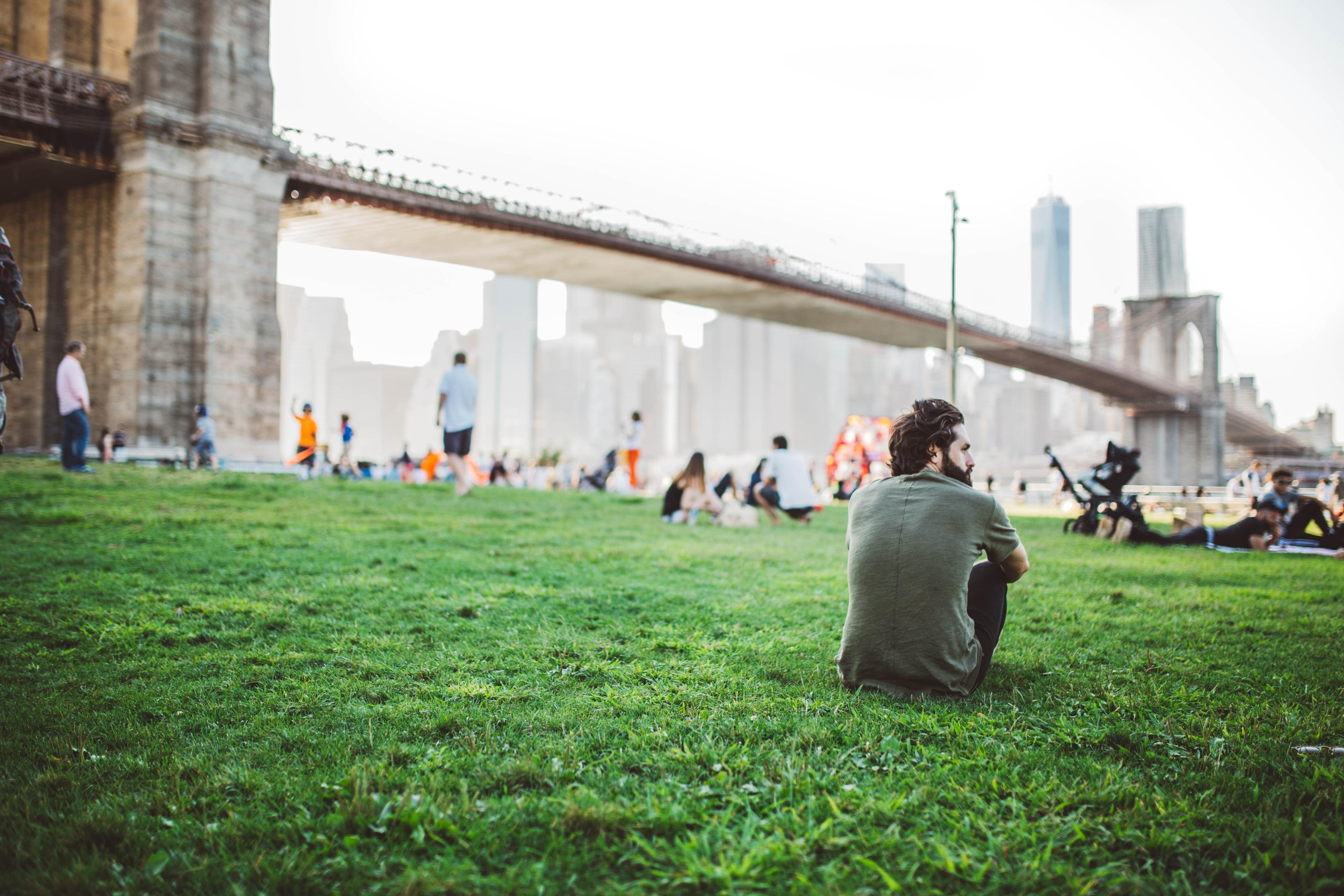 Photo of a man sitting on grass near a bridge with people relaxing and walking in a park setting.