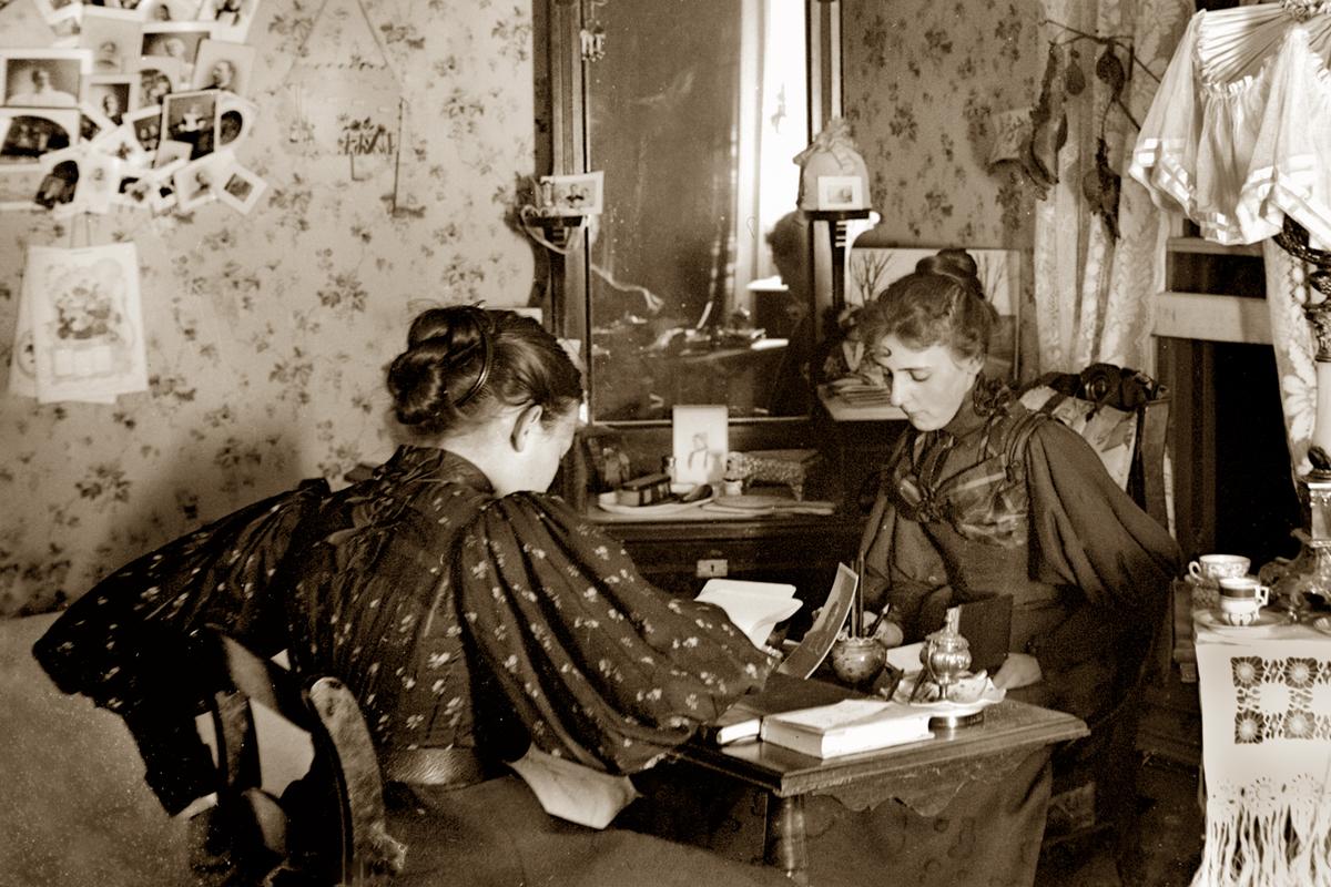 Vintage sepia photo of two women in Victorian attire sitting in a parlour room with floral wallpaper, reading opposite each other at writing desk.