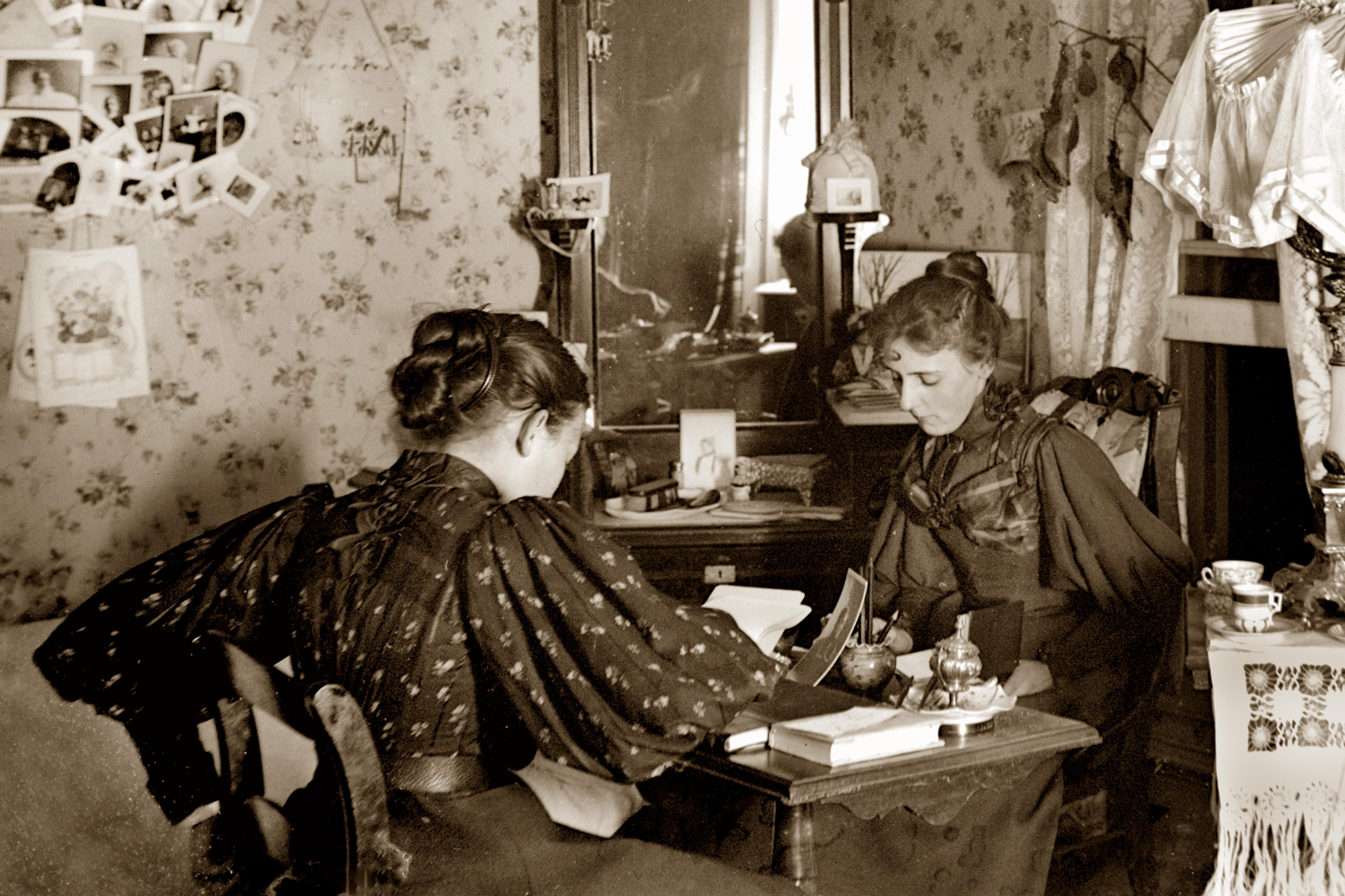 Vintage sepia photo of two women in Victorian attire sitting in a parlour room with floral wallpaper, reading opposite each other at writing desk.