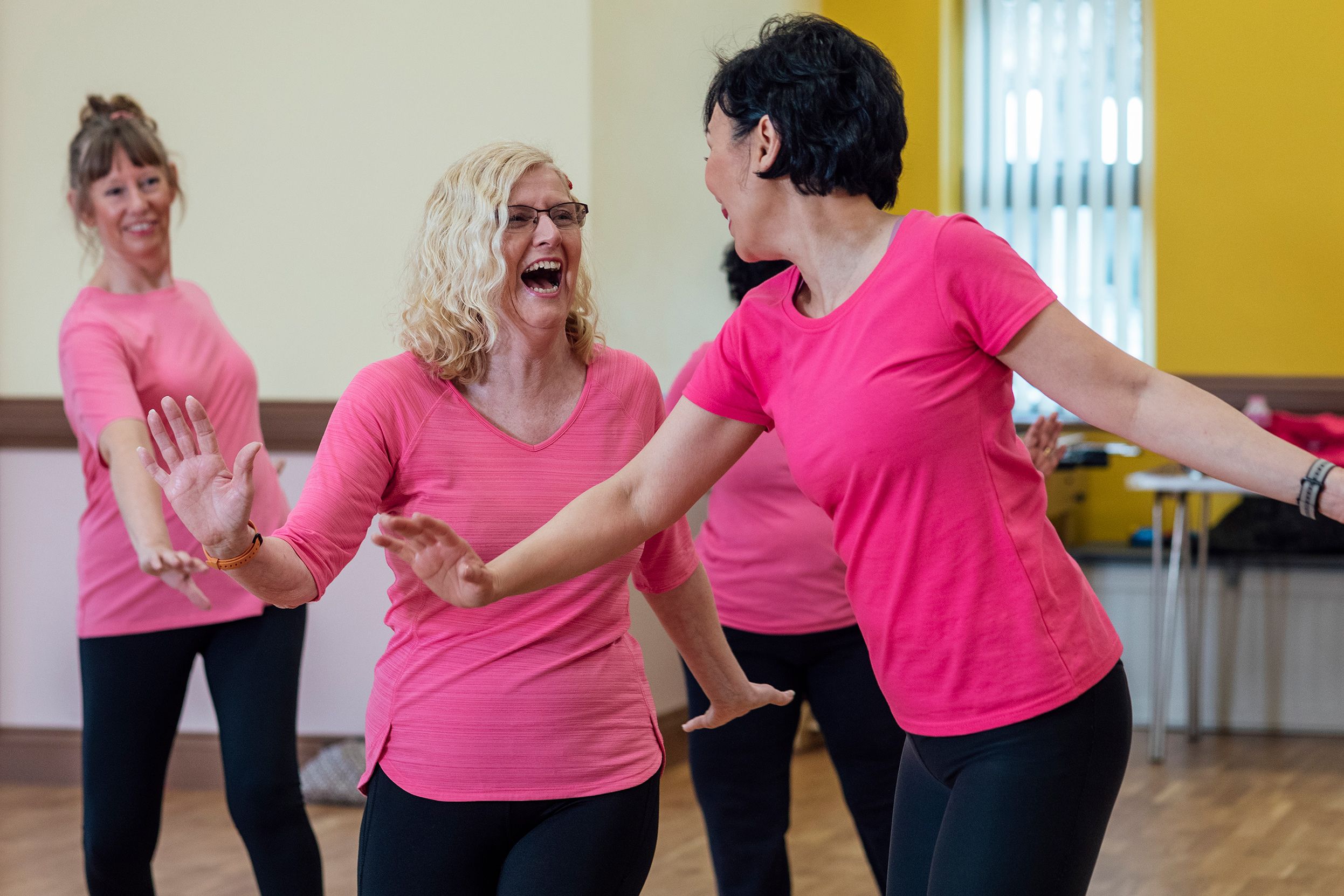 Women laughing and dancing in a studio, all wearing pink tops and black leggings.