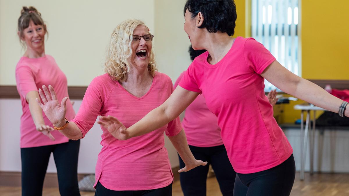 Women laughing and dancing in a studio, all wearing pink tops and black leggings.