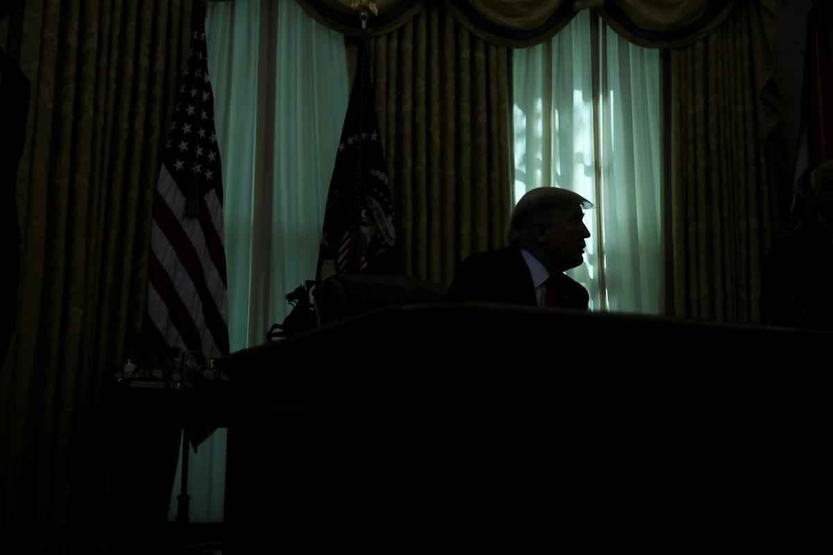 Silhouetted figure at a desk with US flag nearby, backlit by sunlight through curtains.