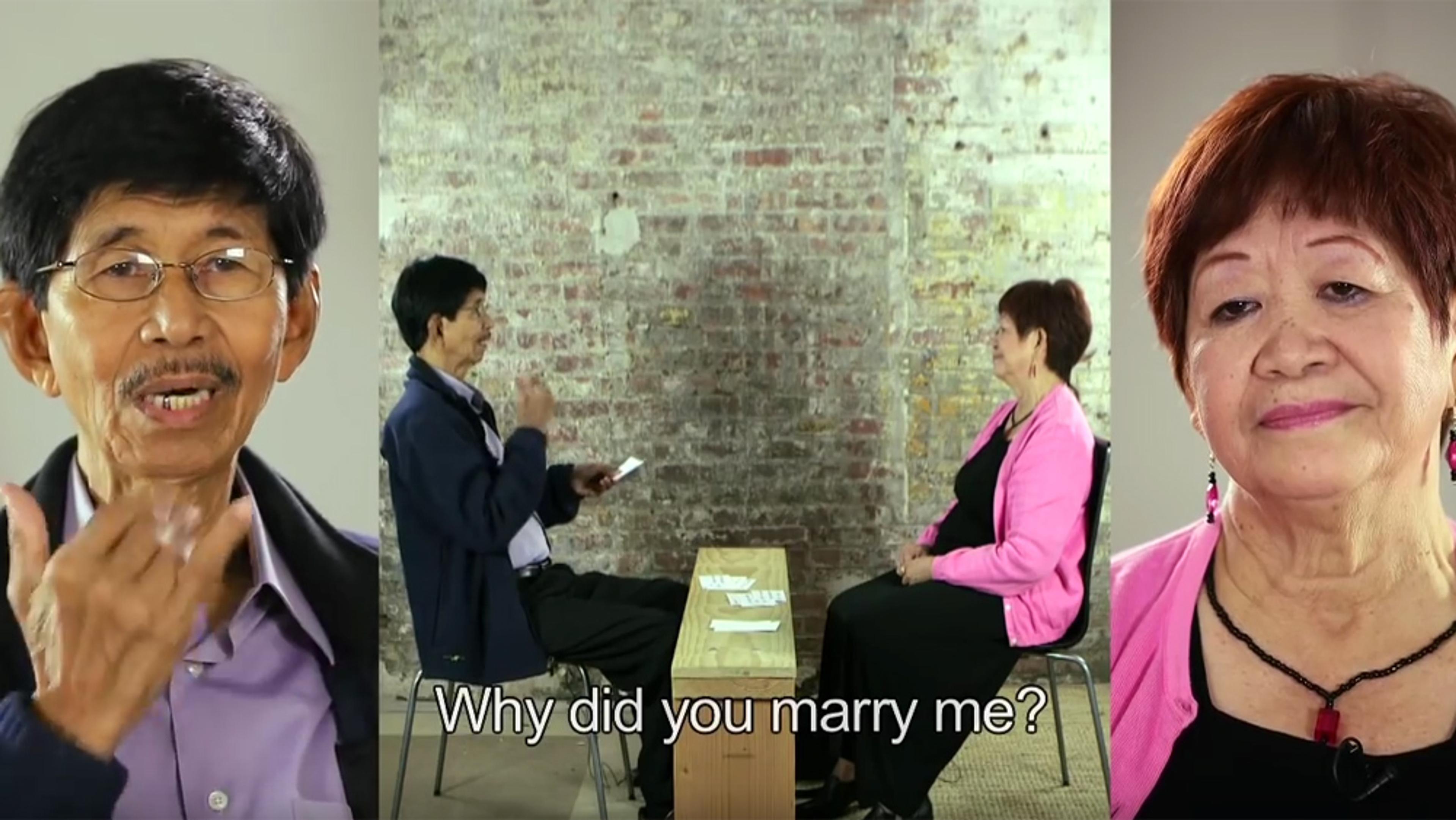 Photo of an elderly couple facing each other on a stage and speaking. Text overlay reads ‘Why did you marry me?’ Background wall is textured.