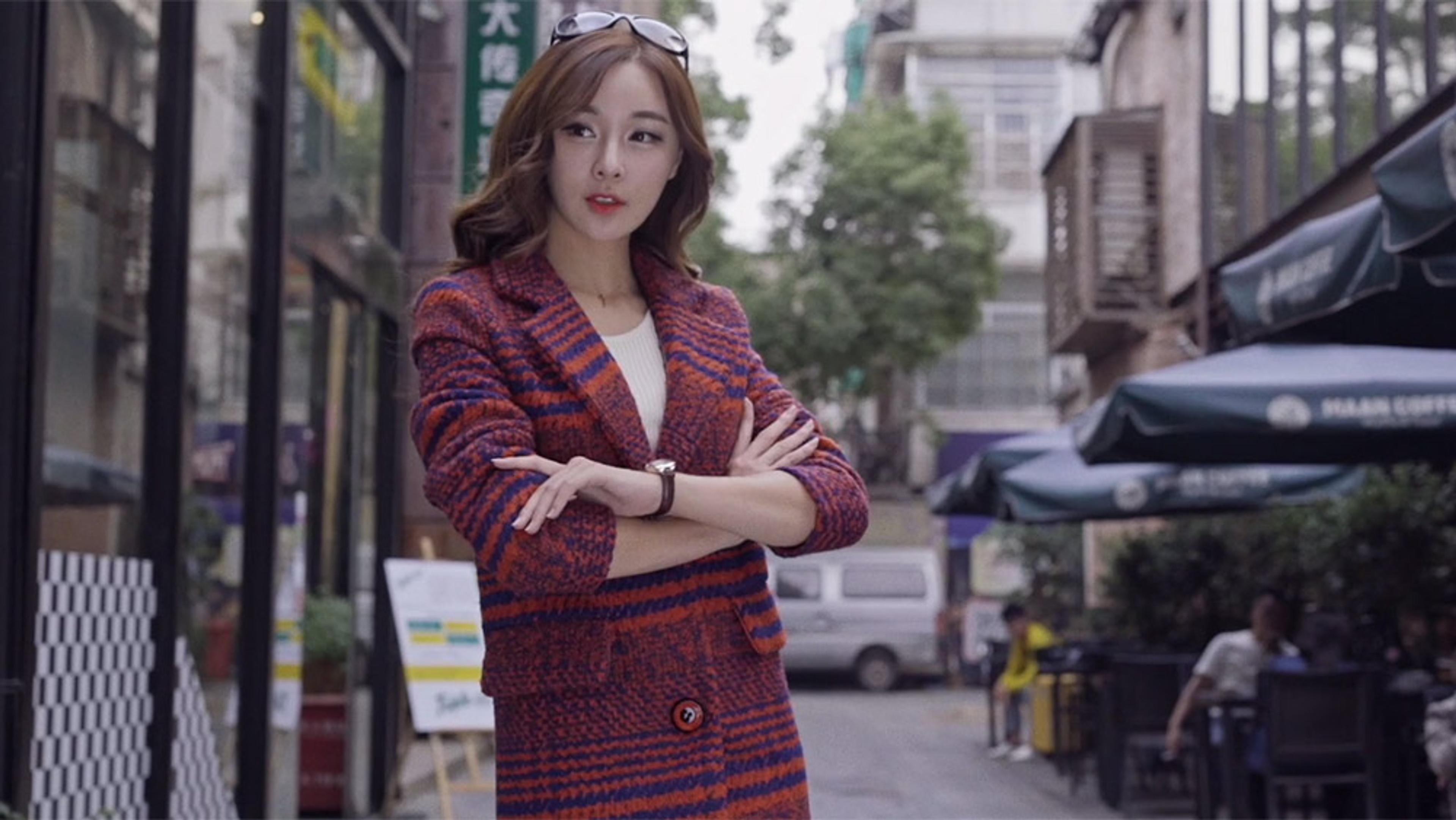 A woman wearing a red and purple coat with arms crossed, standing on a city street lined with shops and trees.