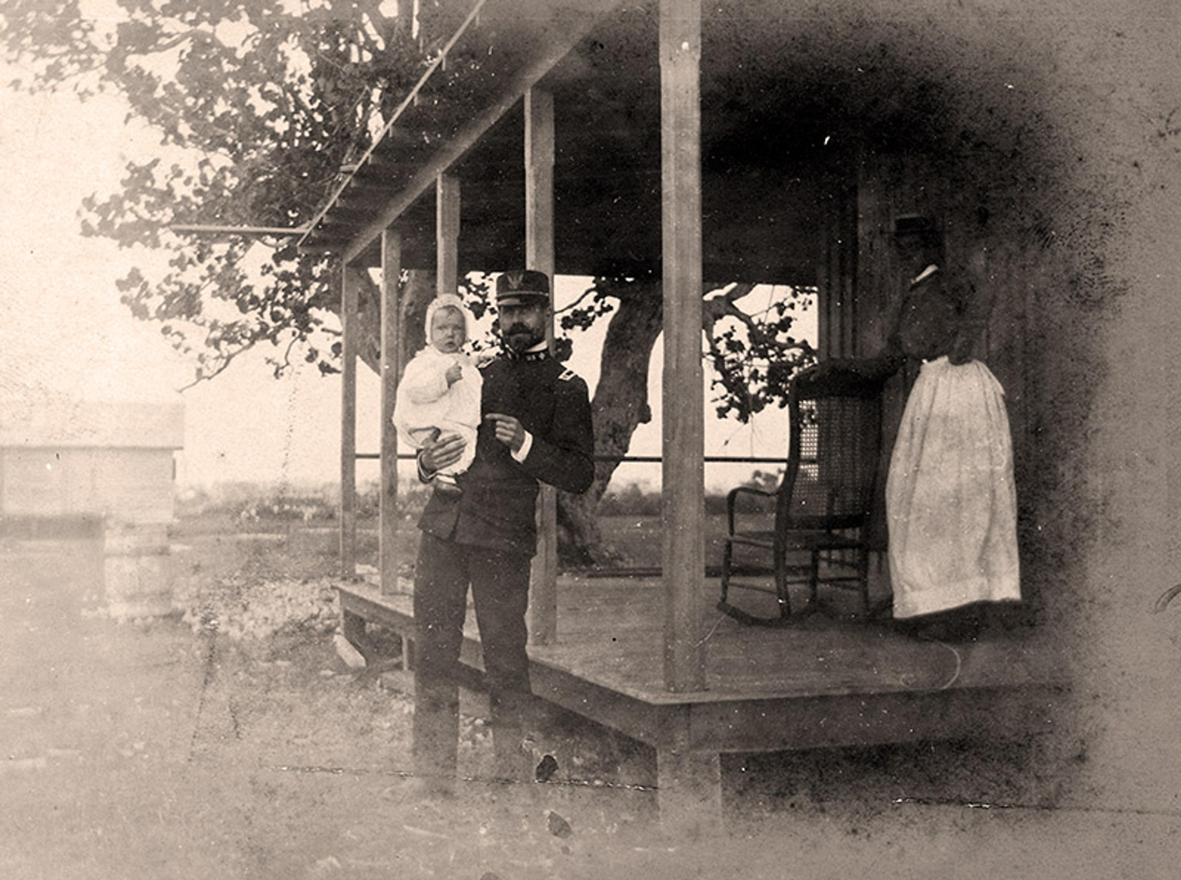 A soldier in uniform holds a young child in his arms beside a veranda