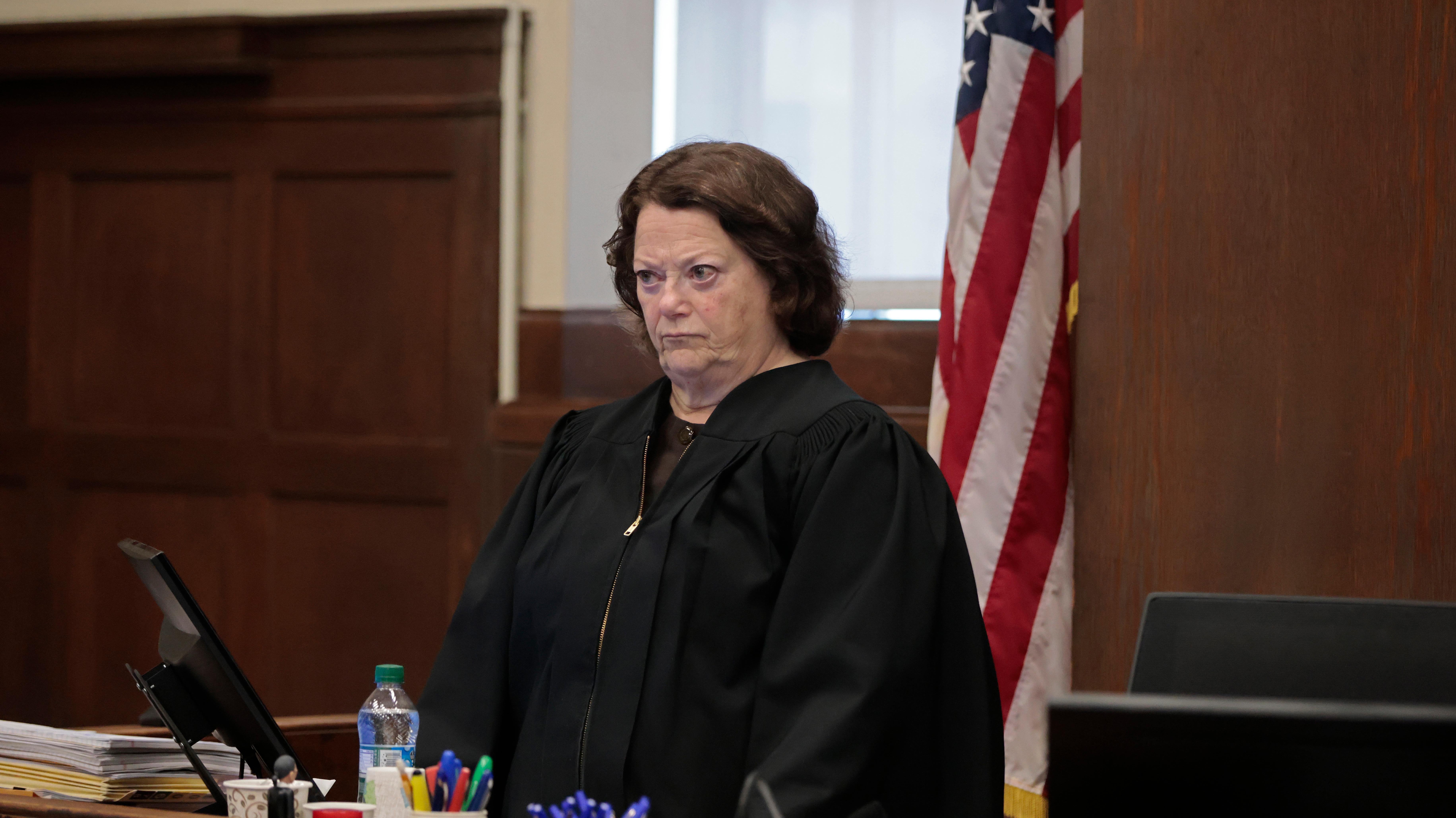 A judge with a stern expression in a courtroom wearing a black robe standing in front of a US flag.