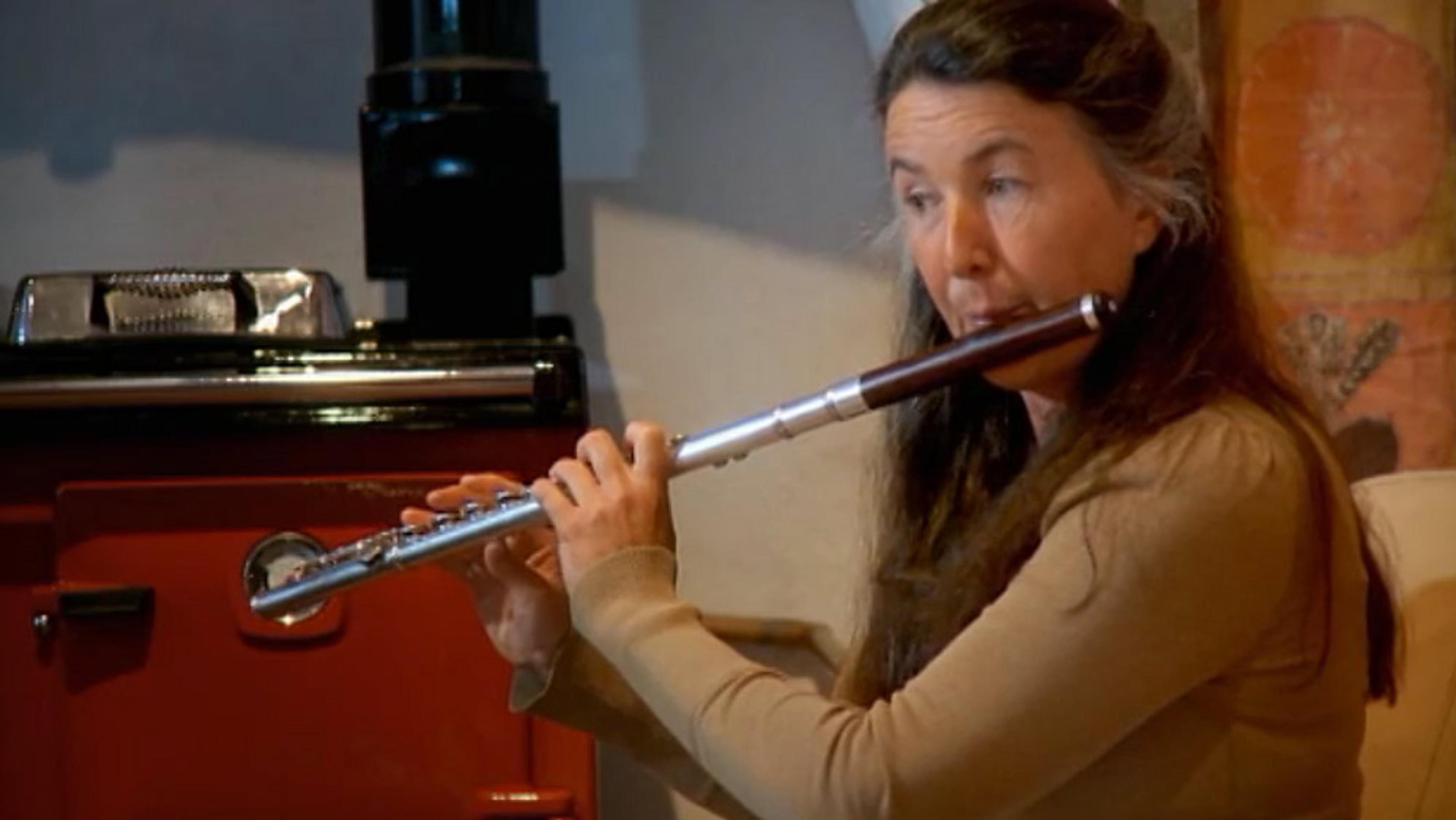 A woman with long dark hair and focused expression playing a flute next to a red stove in a home setting.