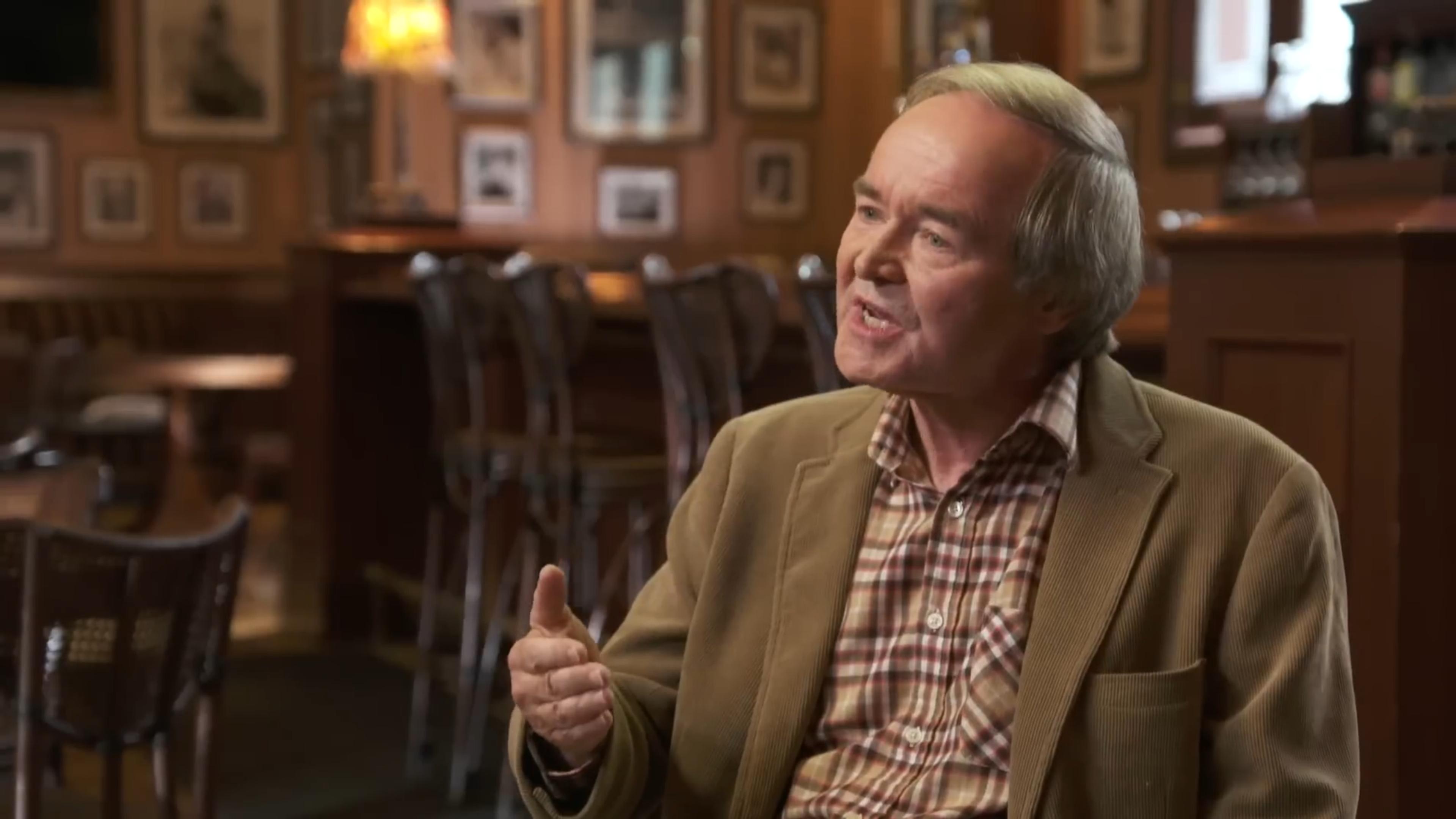 A man speaking in a warmly lit pub with vintage decor in the background.