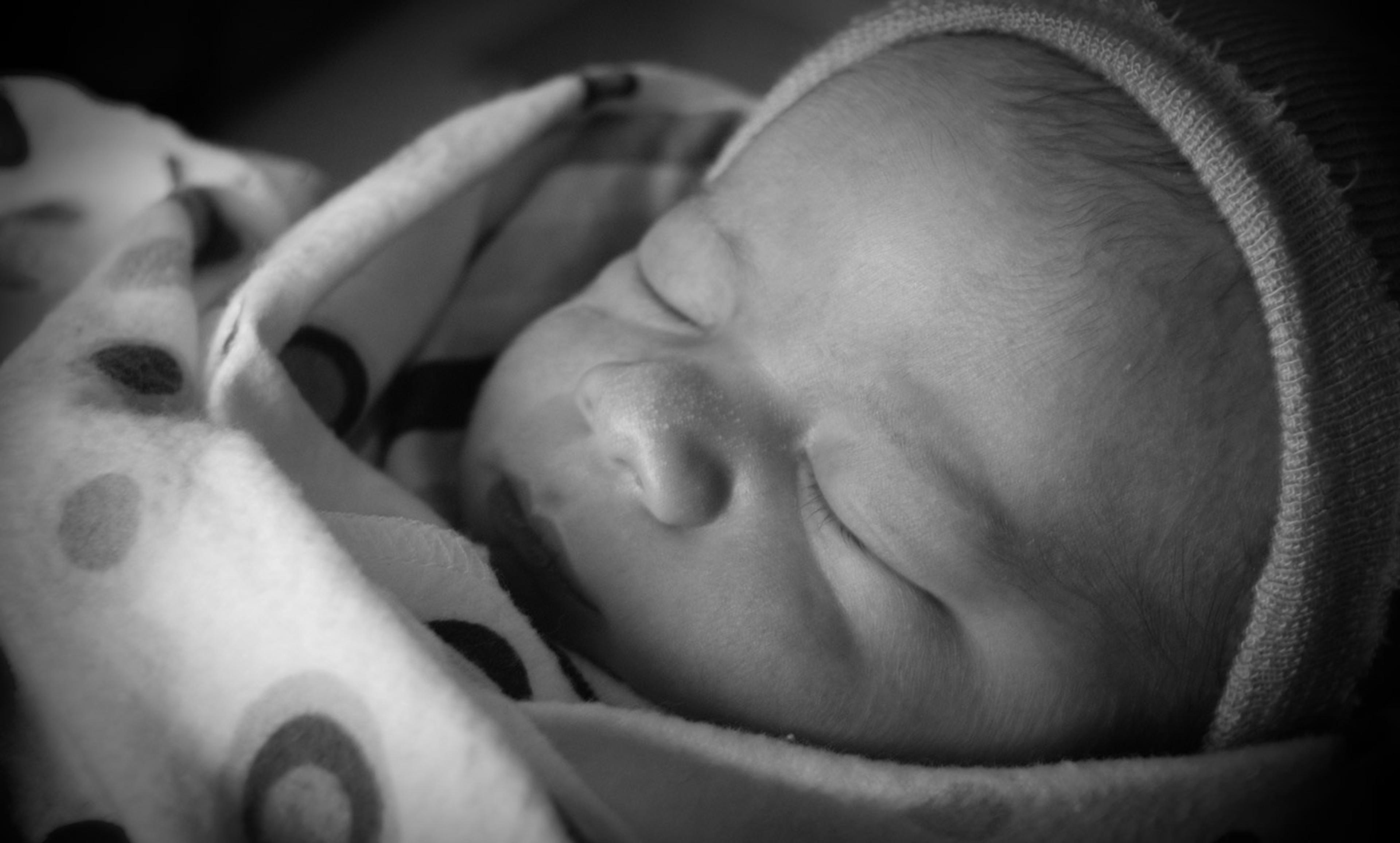 Black and white photo of a sleeping baby wearing a beanie and wrapped in a polka dot blanket.