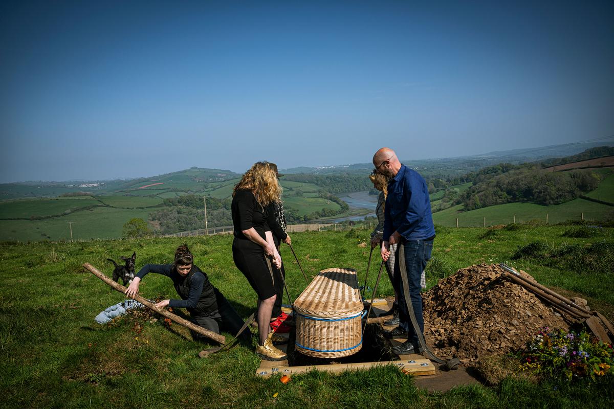 People lowering a wicker coffin into a grave in a green landscape with hills and a river in the background.