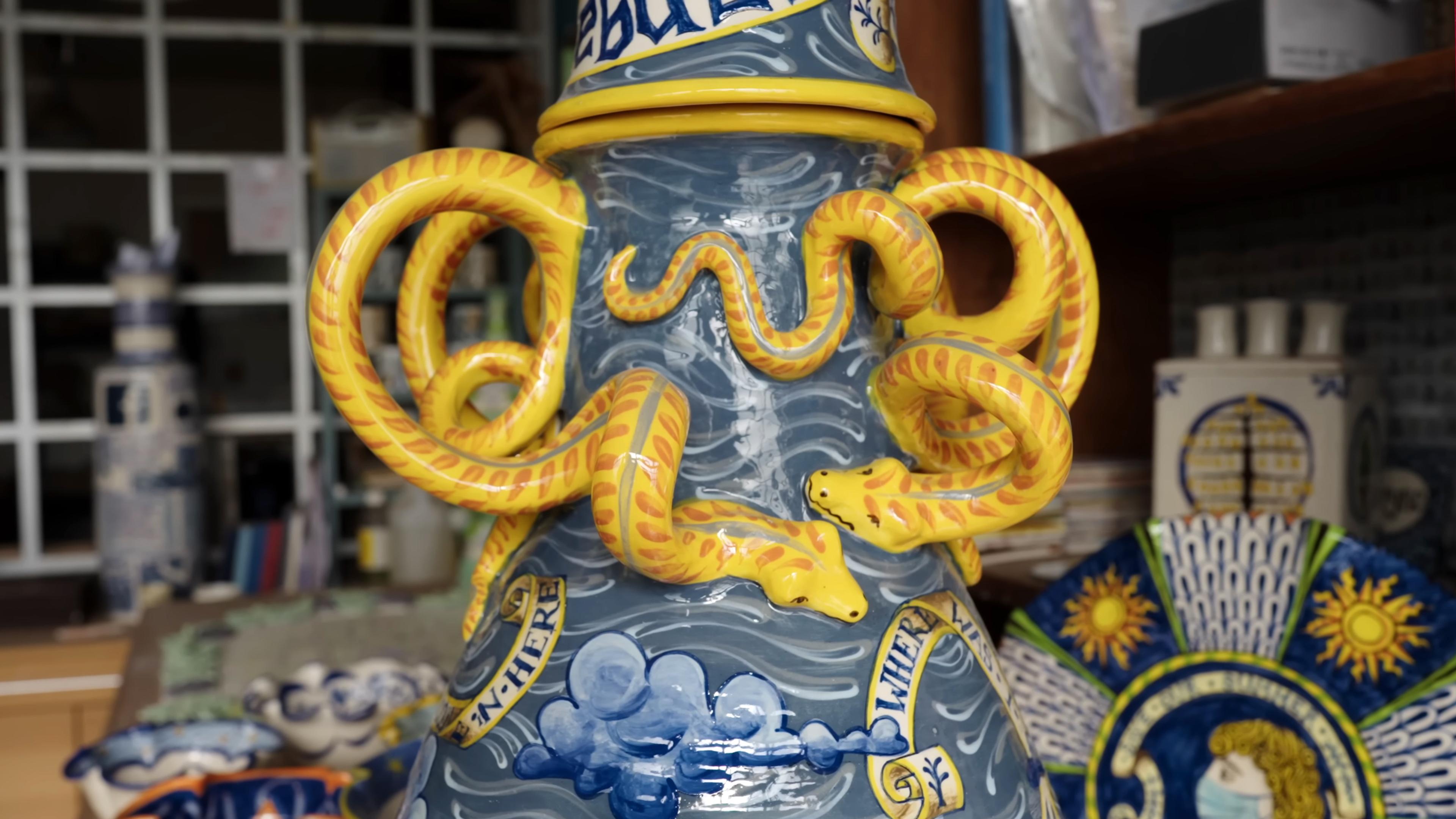 Ceramic vase with yellow snake handles blue clouds and text detail in a workshop setting.