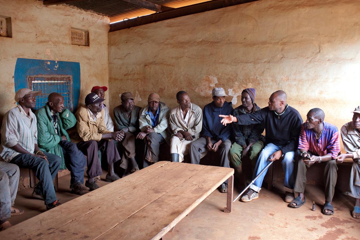 A group of men sitting along a wall in a rustic room, engaging in conversation with visible gestures.
