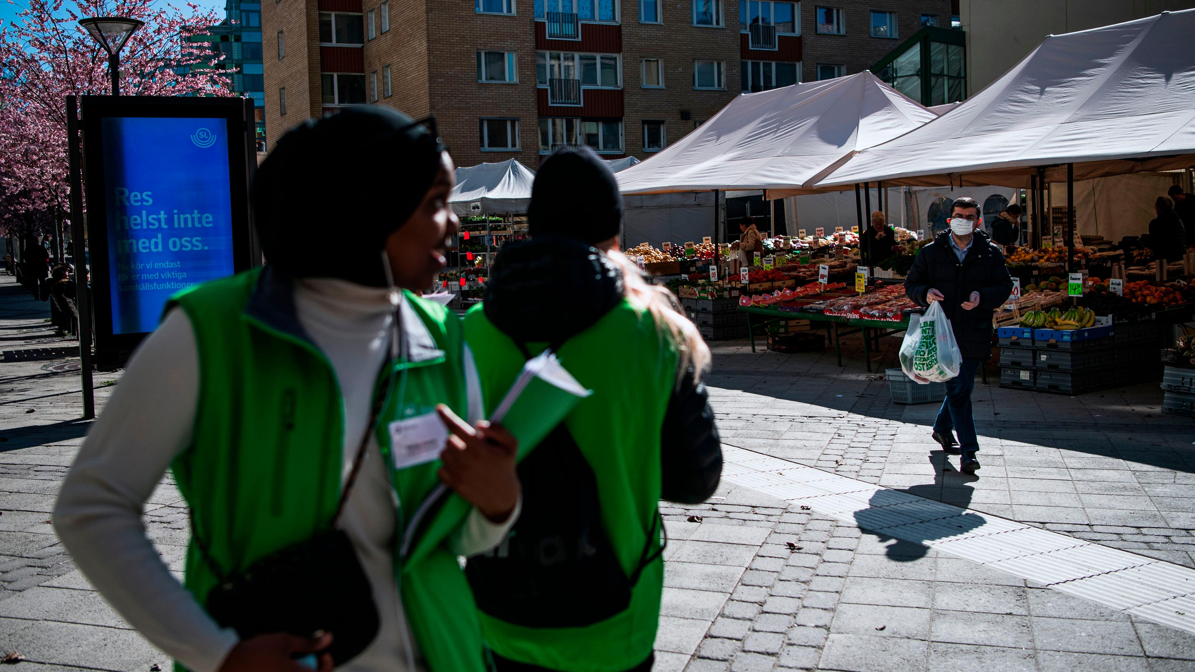 Photo of an outdoor market with fruit stalls, a masked man carrying bags and two people in green vests walking past.
